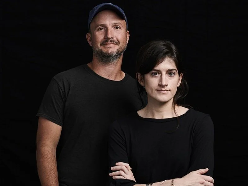 A man with a beard and a blue cap standing next to a woman with arms crossed, both wearing black shirts, against a black background.