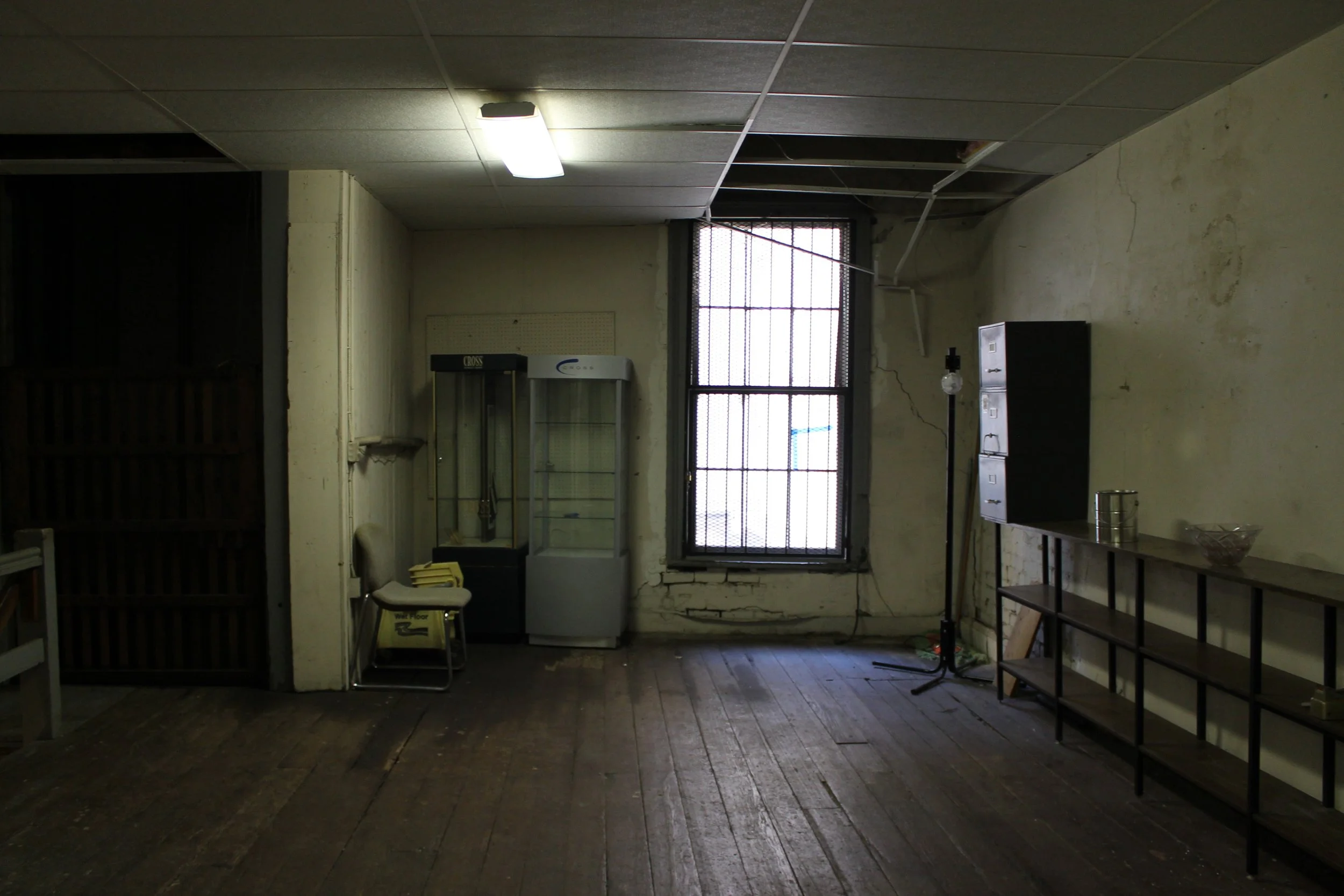 Interior of an old, dimly lit room with worn hardwood floors, peeling paint on walls, a window with bars, a shelving unit, a chair, a vending machine, a lamp, a closed cabinet, and some small items on the shelves.