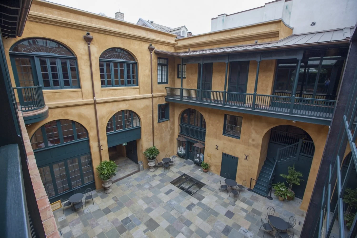 Indoor courtyard with yellow stucco walls, large arched windows, and green railings on the second floor. The ground level has tables and chairs, potted plants, and a fountain. There are outdoor stairs on the right side leading to the upper level.