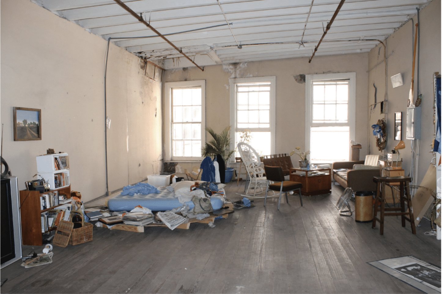 Messy living room with scattered clothes, books, and clutter on the floor, with furniture including sofas, chairs, and a coffee table, and three large windows letting in natural light.