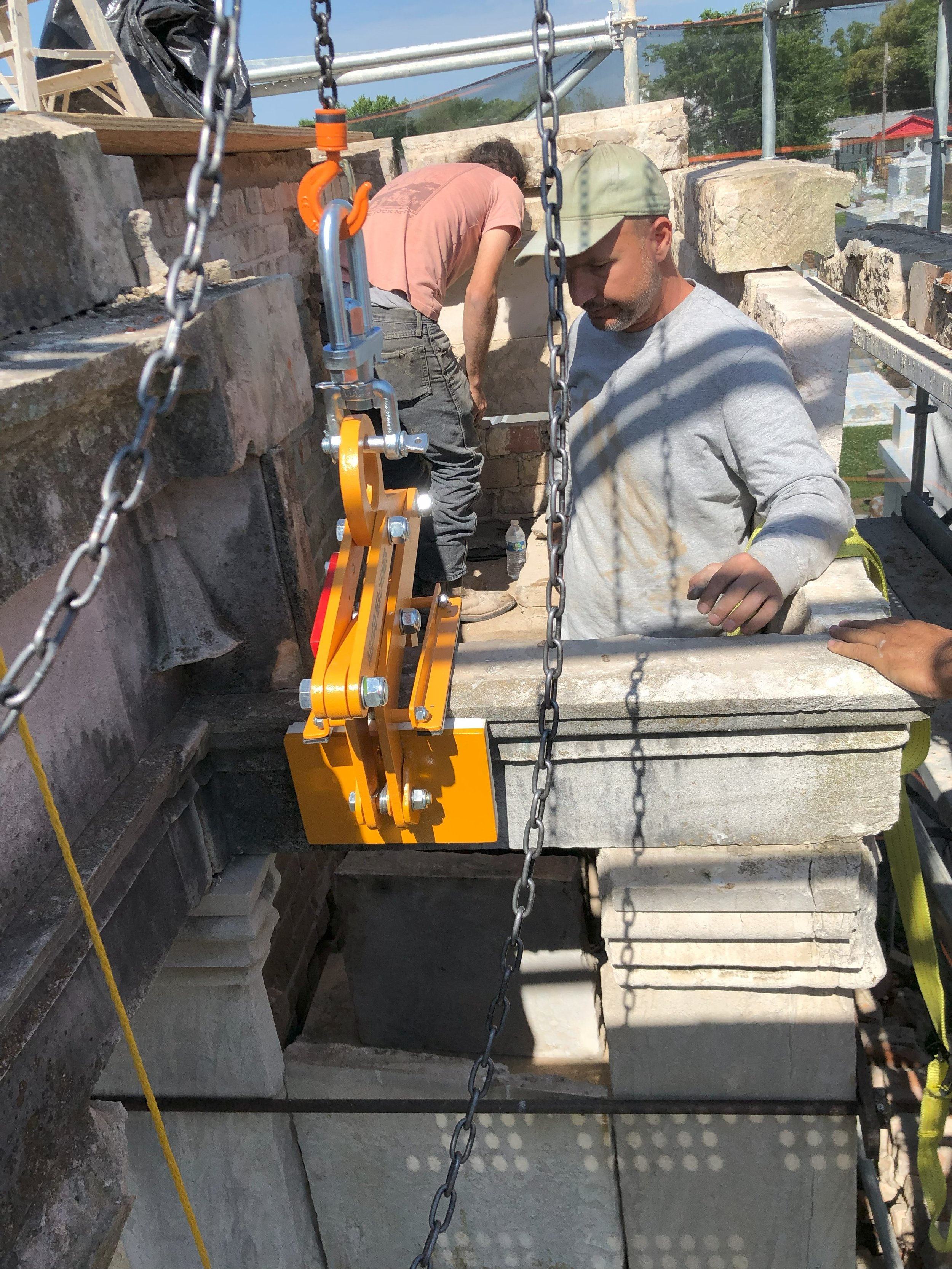 Two construction workers are on a scaffolding installing or repairing stone bricks or tiles. One worker, wearing a gray shirt and a camo cap, is focused, while the other person in a pink shirt is bending over in the background. Safety chains and construction equipment are visible.