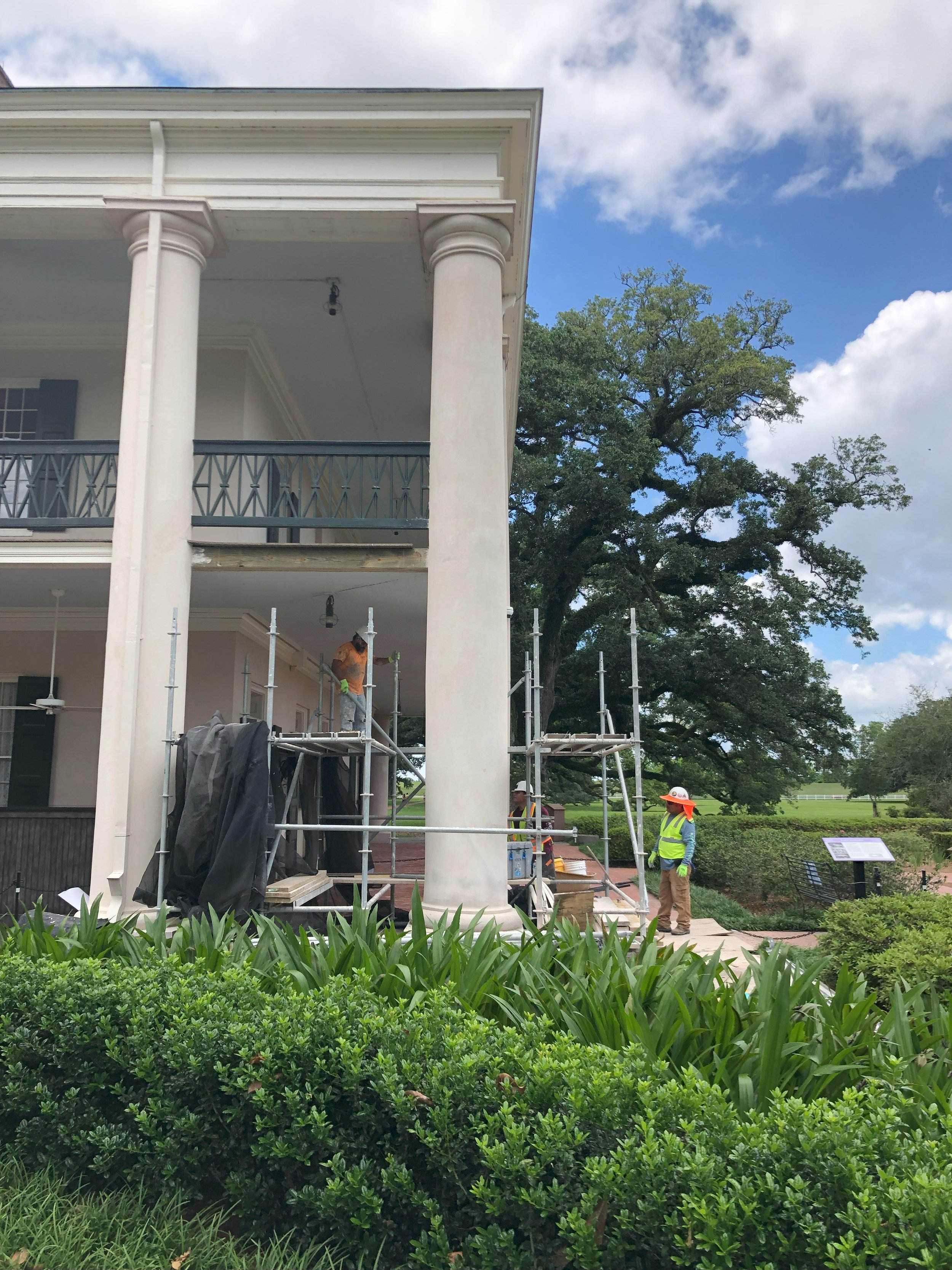 Construction workers on scaffolding outside a white, two-story building with large columns, green lawn, and trees.