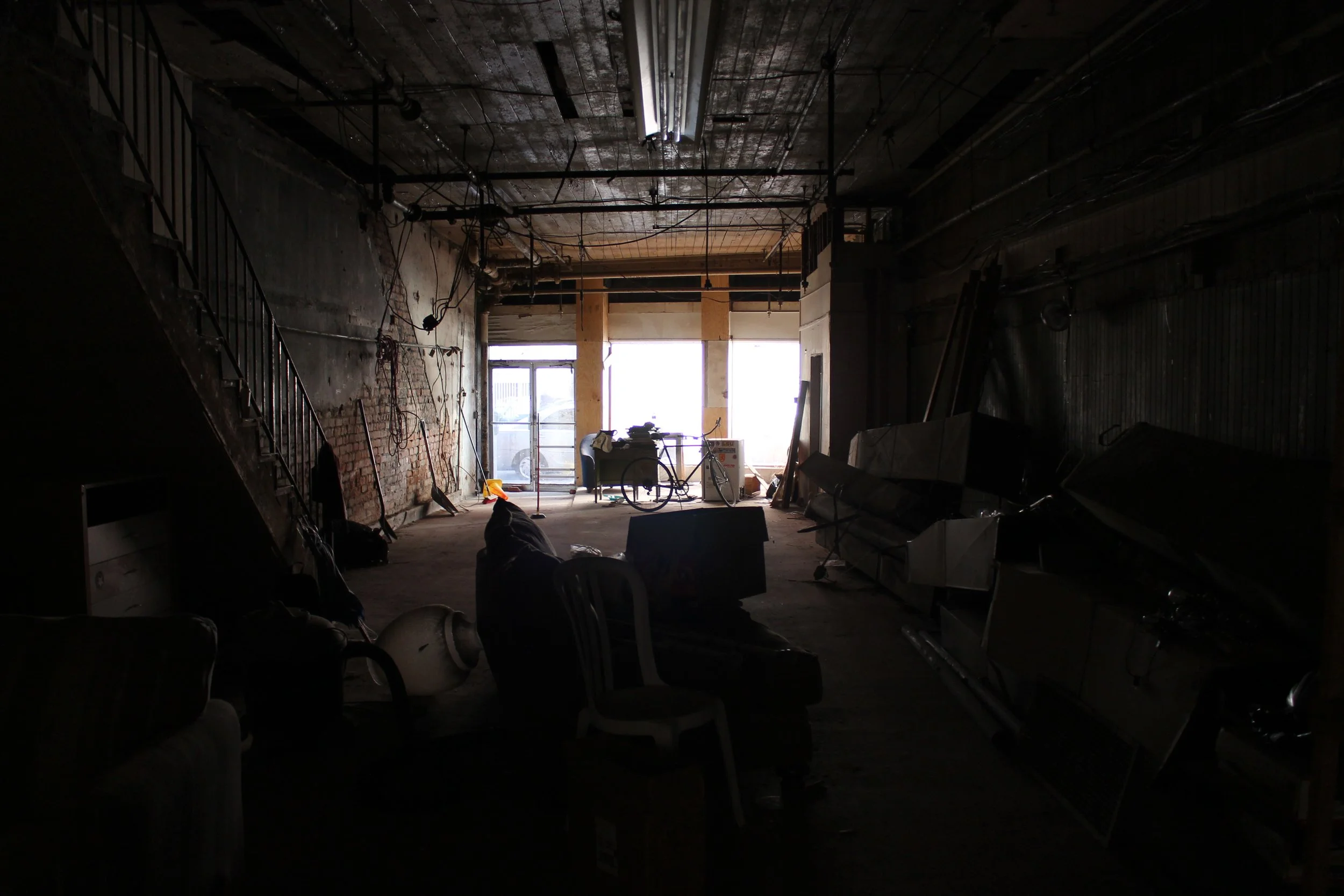 Interior of a dimly lit, cluttered room with exposed brick wall, ladder, and construction materials, illuminated by sunlight coming through large windows.