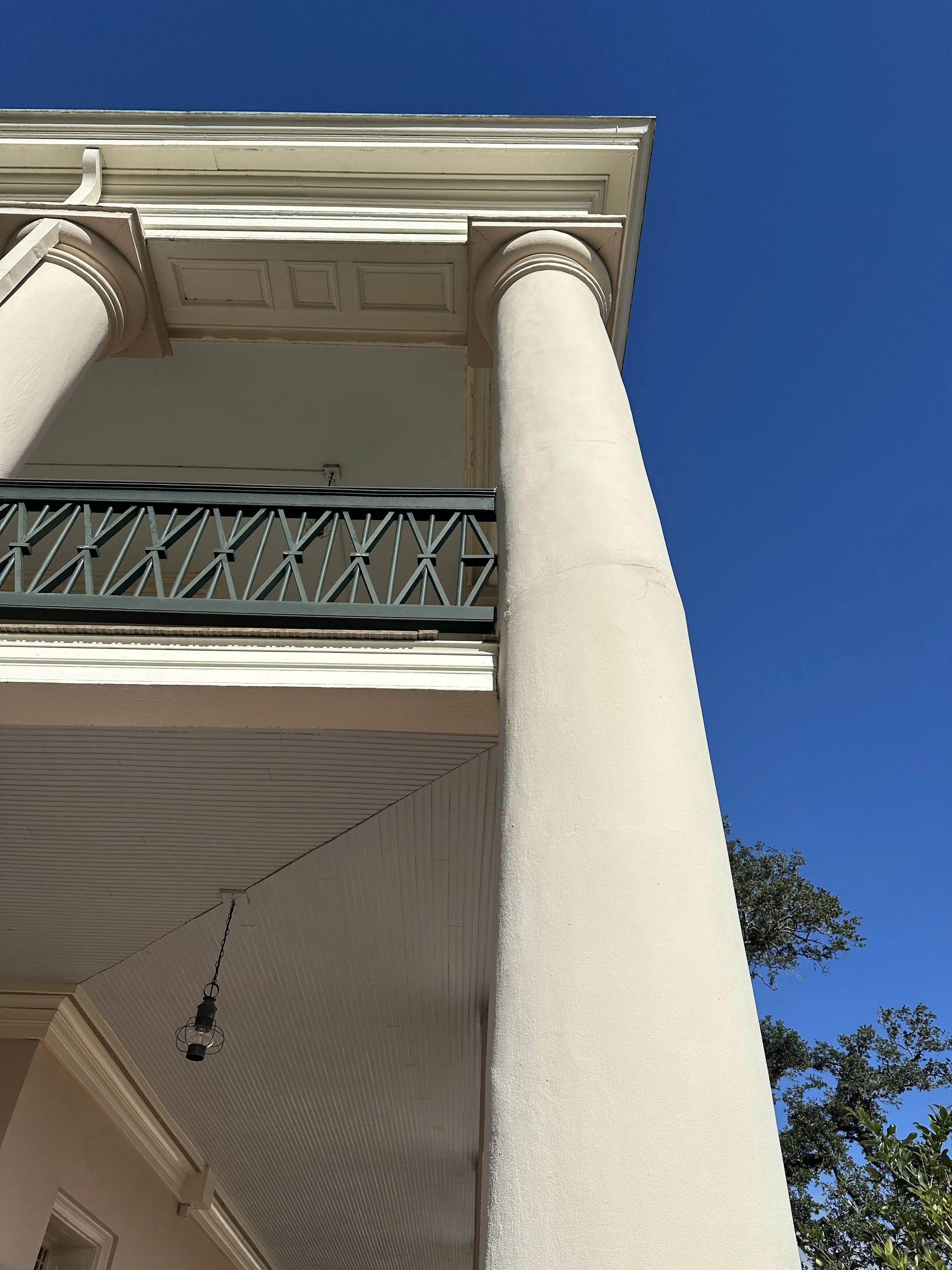 Close-up of a white building with a large decorative column and a balcony with a black railing, set against a bright blue sky.