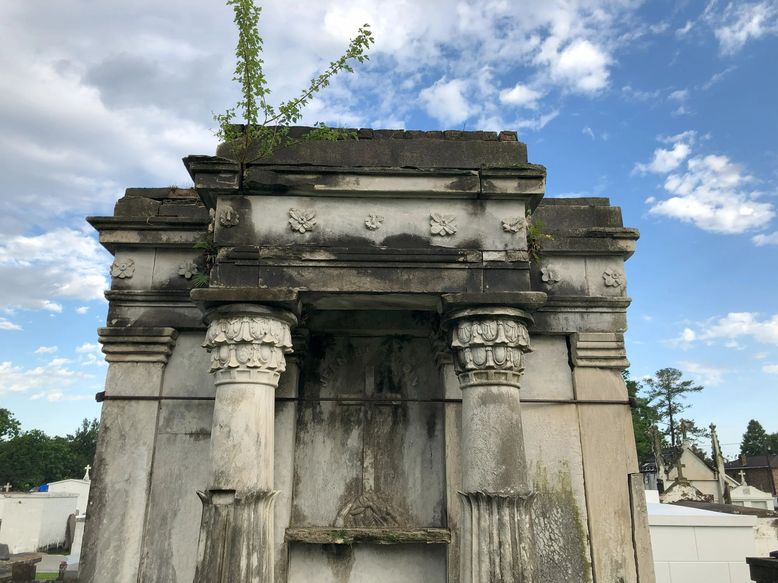Old, weathered mausoleum or monument with carved columns and floral designs, in a cemetery with trees and other tombstones in the background, under a partly cloudy sky.