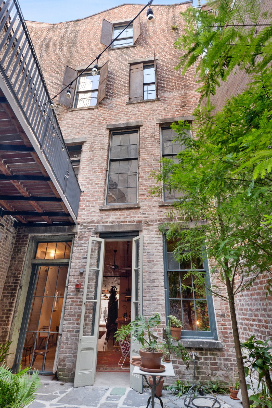 A brick townhouse with multiple windows, some open, and a small outdoor patio area with potted plants, a table, and a string of lights hanging above. New Orleans Courtyard Cypress Building Conservation masonry restoration and consulting