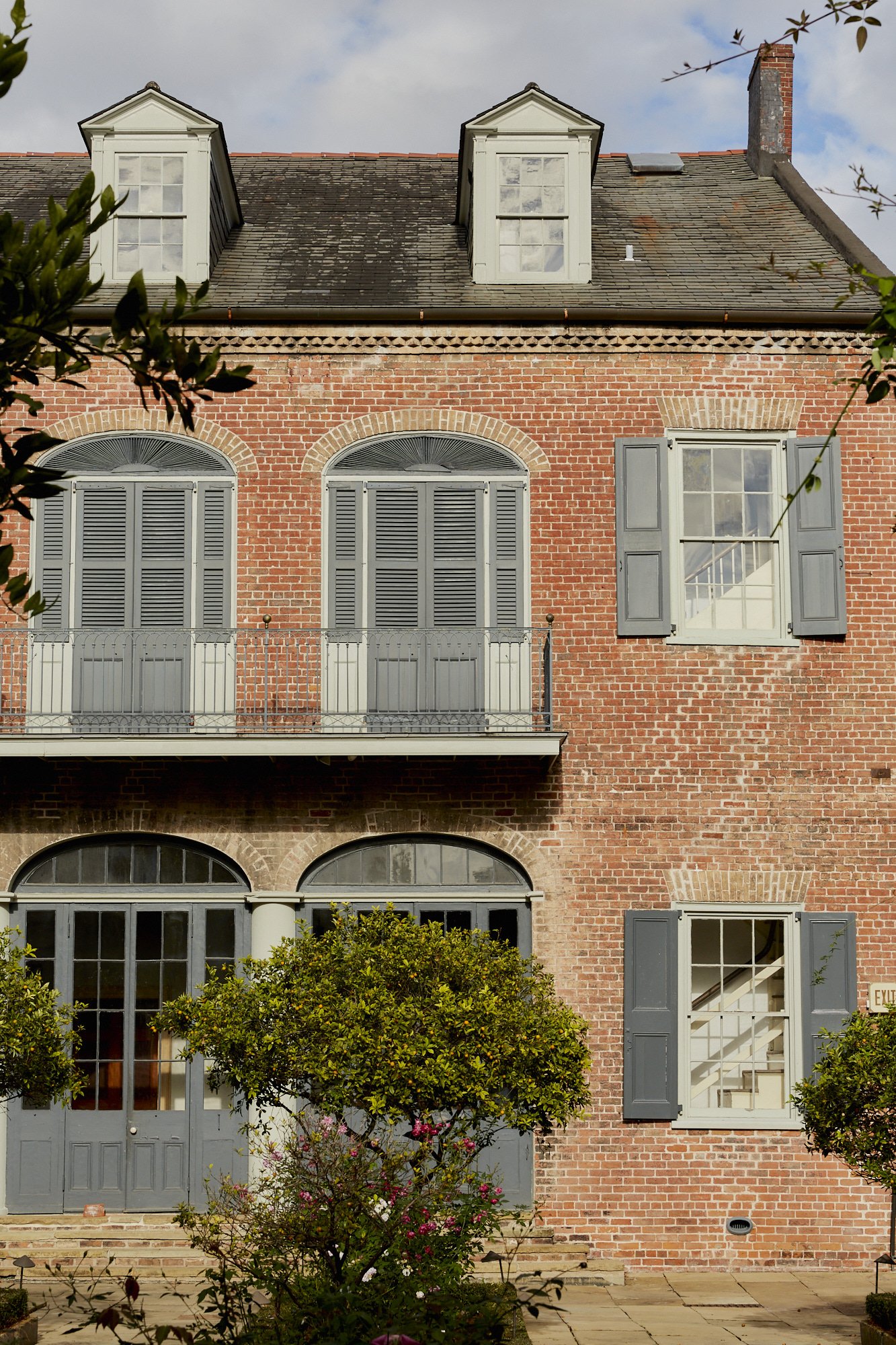 A multi-story red brick building with gray shutters on the windows, some open, and white window frames. There are trees and bushes in front of the building, with a partly cloudy sky above.