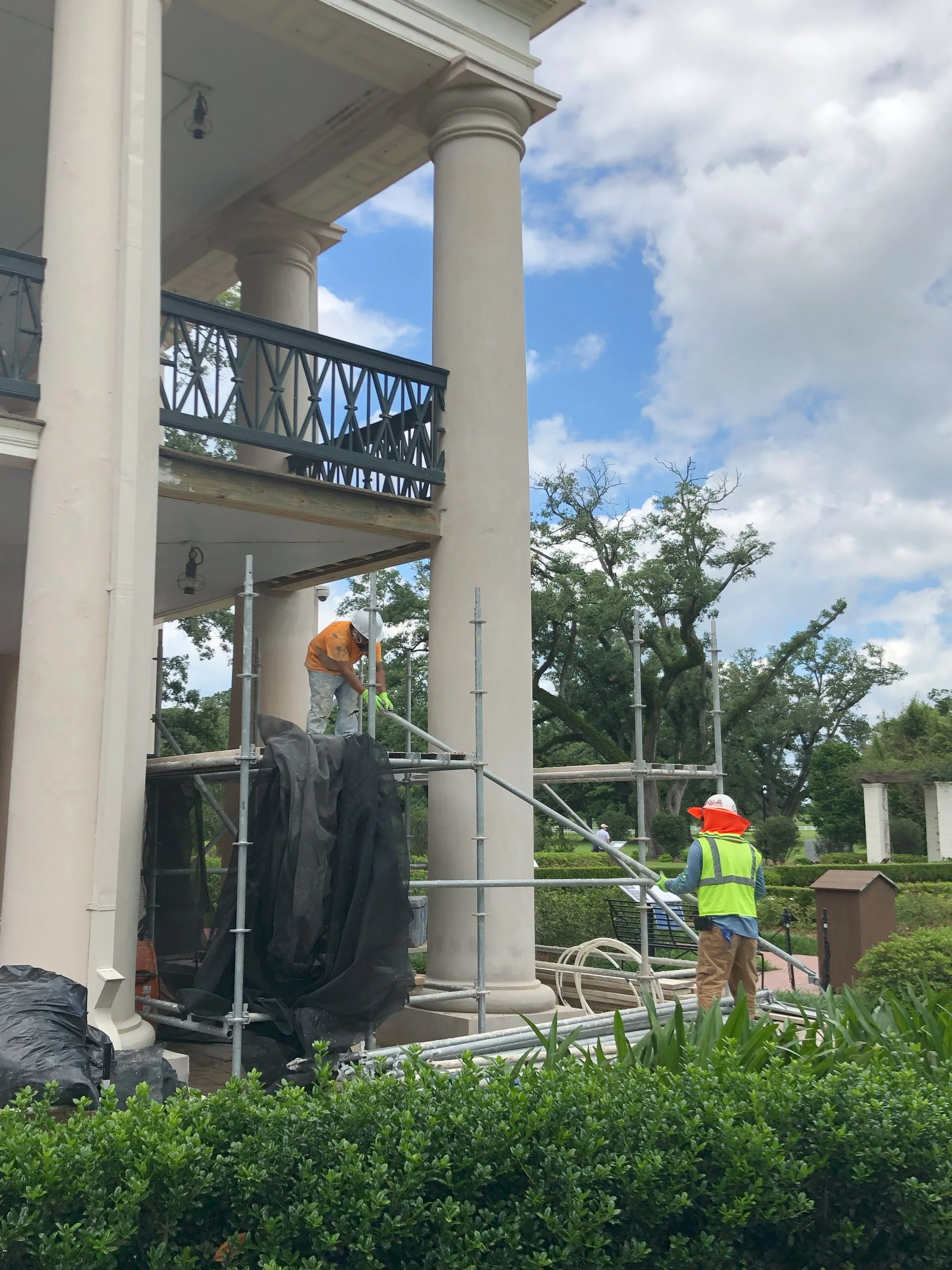 Construction workers cleaning or inspecting scaffolding on the porch of a large, classical style building with columns and a balcony, with trees and a cloudy sky in the background.