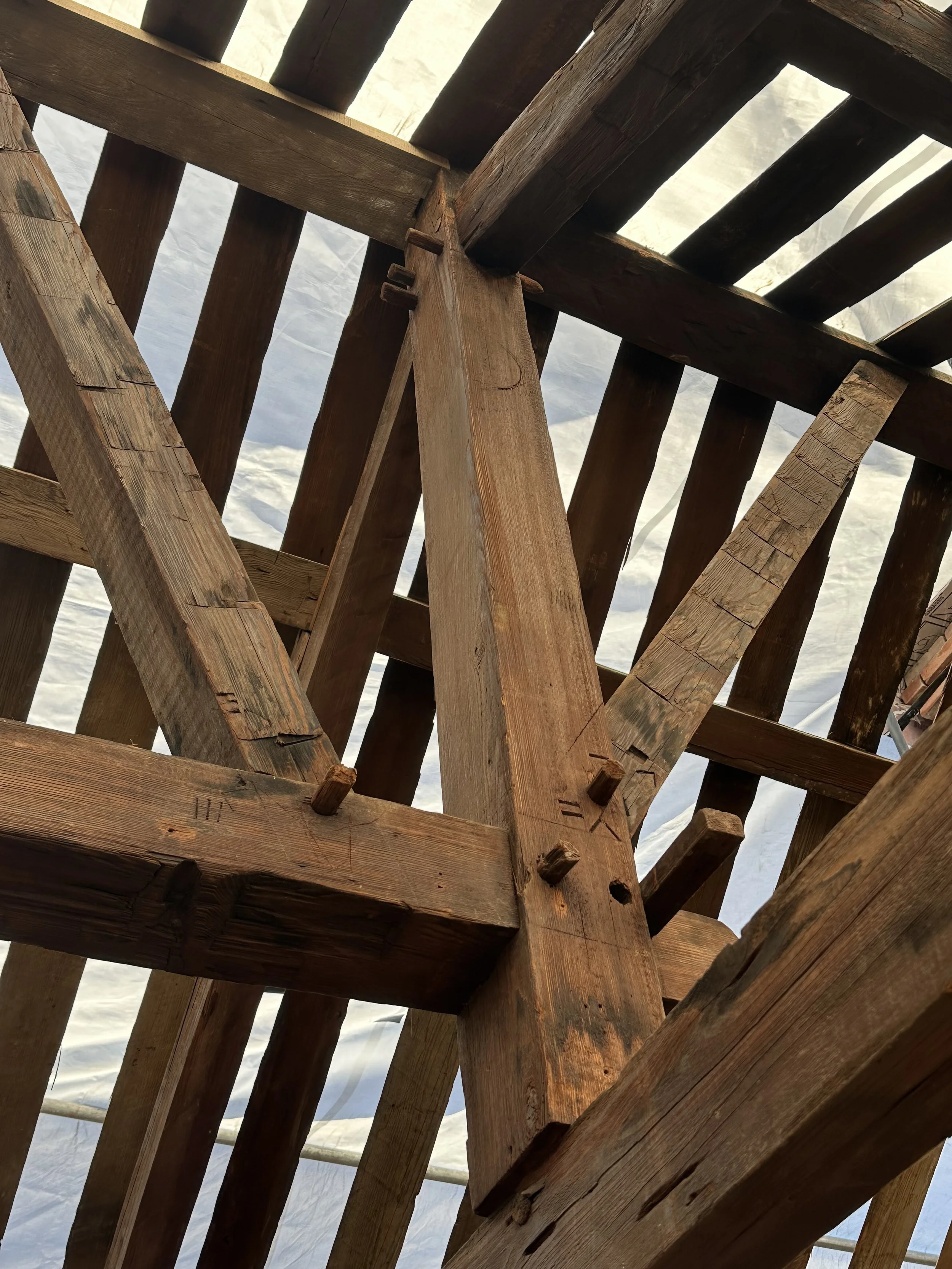 Close-up view of a wooden framework or structure, showing intersecting beams and nails, with the sky and clouds visible in the background.