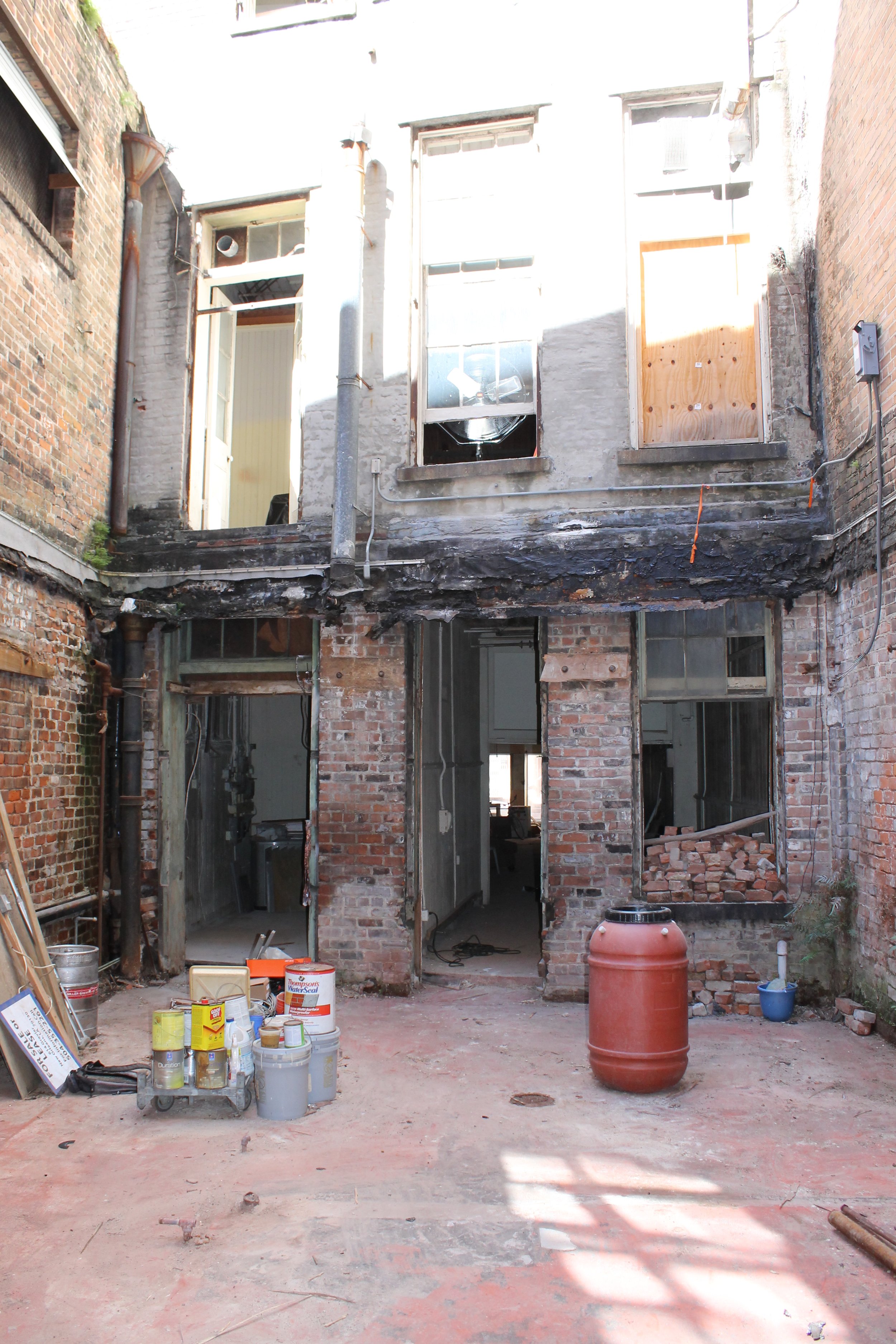 An outdoor view of a brick building undergoing renovation, with construction supplies and tools on the ground, and two windows and a door on the upper level, some of which are boarded up.