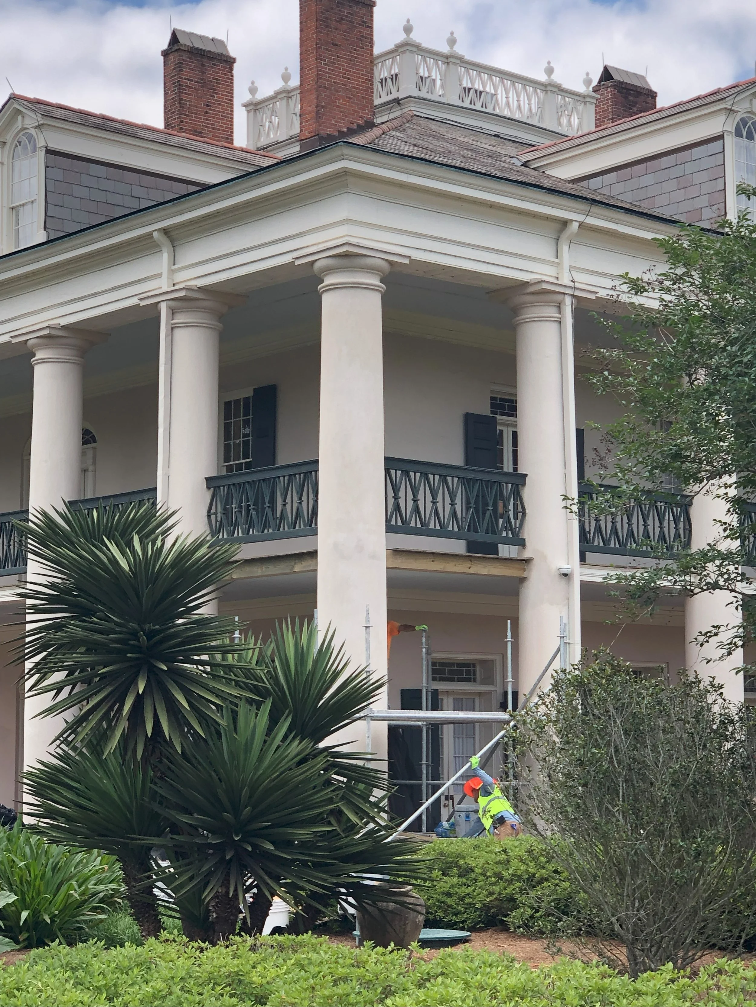 Workers repairing scaffolding on the porch of a large, white, historic house with tall columns and a second-story balcony.