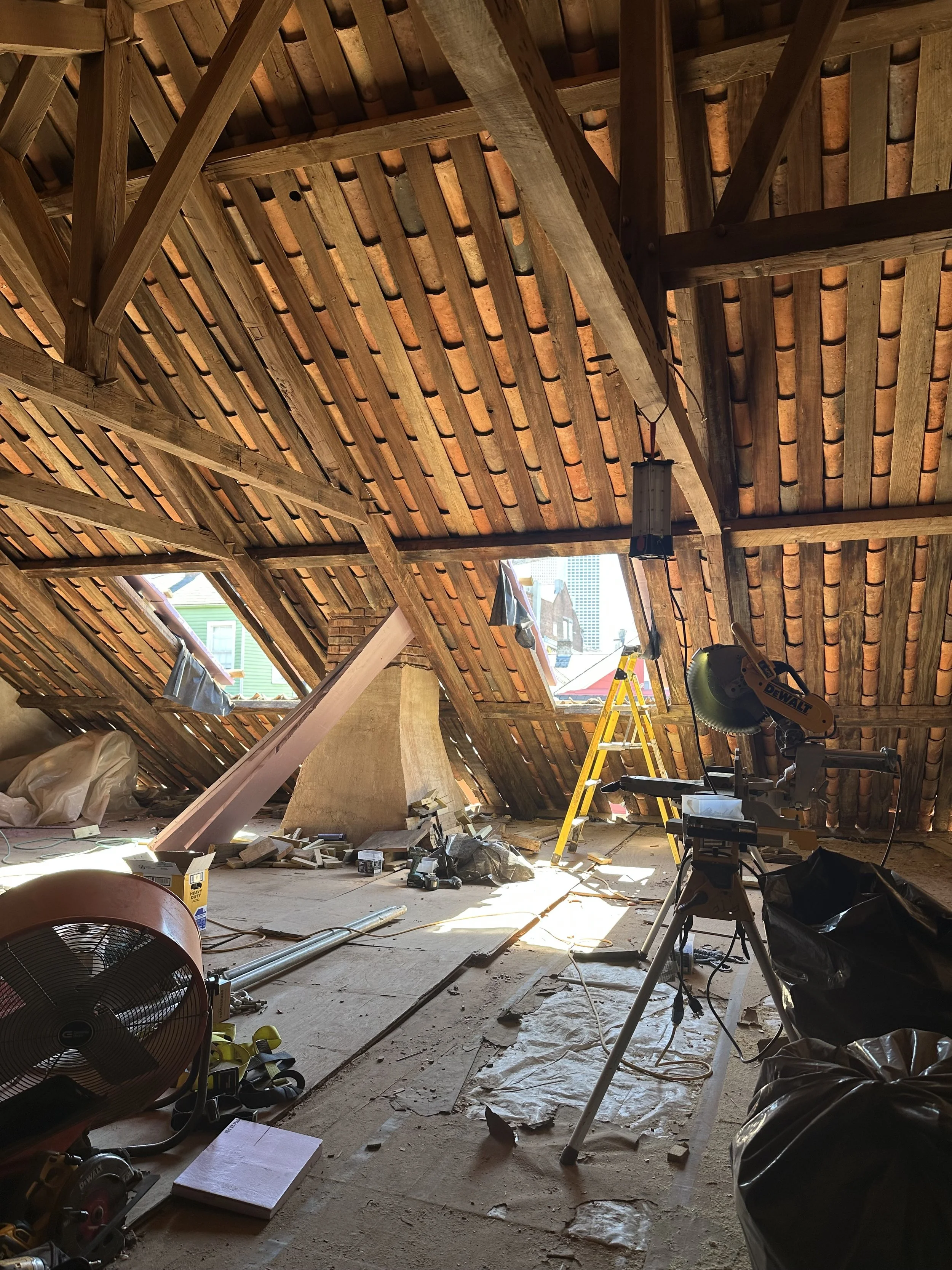 Attic under renovation with roofing structure visible, tools, ladder, and construction materials scattered around.