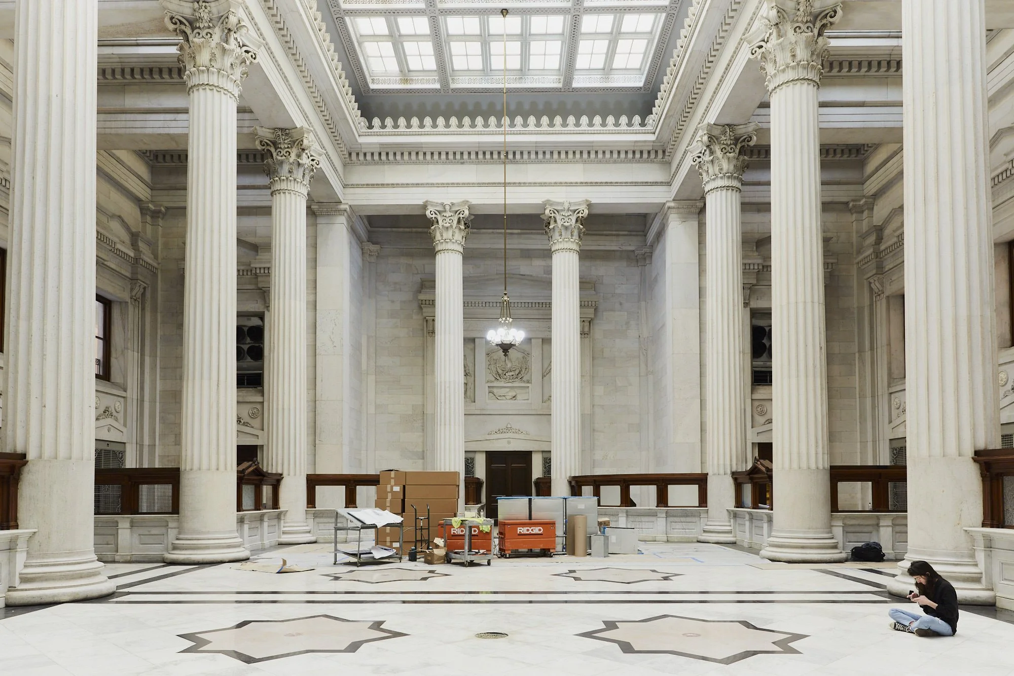 Interior of a grand, classical building hall with tall Corinthian columns and a glass skylight ceiling. There are construction supplies and a person sitting on the floor in the lower right corner.