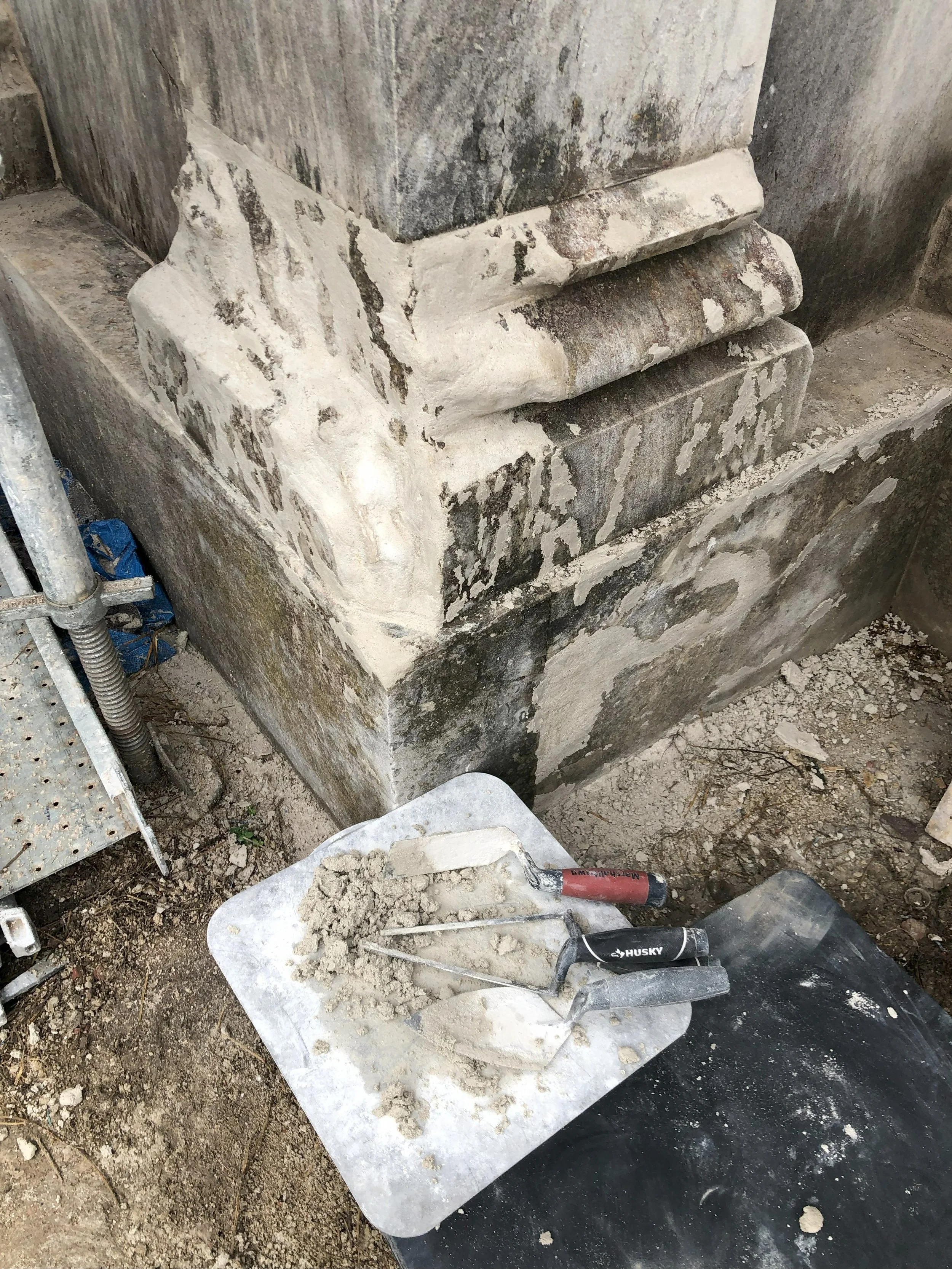 Close-up of an old, weathered stone foundation with chipped paint and exposed concrete, with trowels and a black trowel handle resting on a white tray with mortar nearby, indicating ongoing masonry or restoration work.