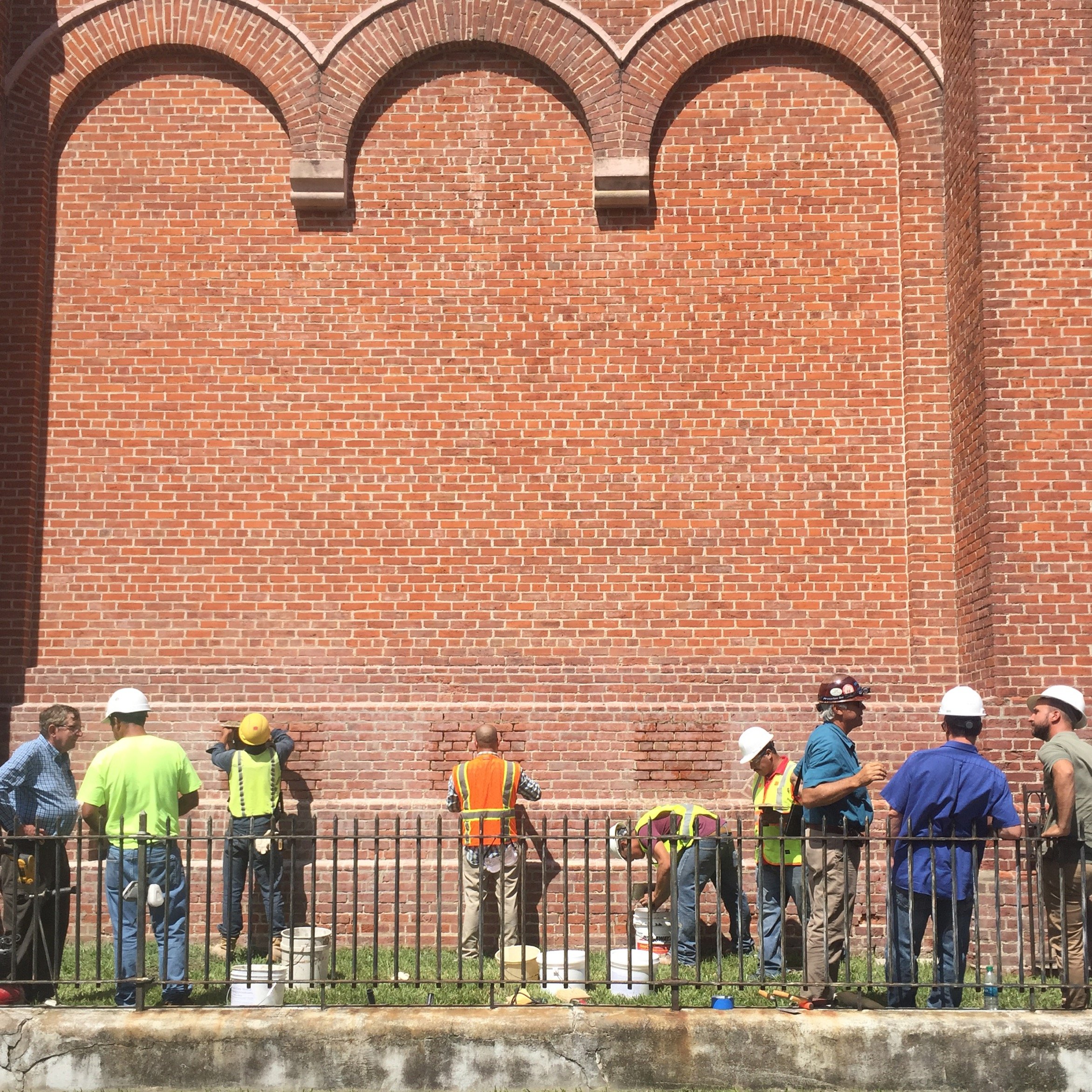 Construction workers and engineers in safety vests and helmets working on a brick wall outside, with a black metal fence in front. Historic masonry services New Orleans. 