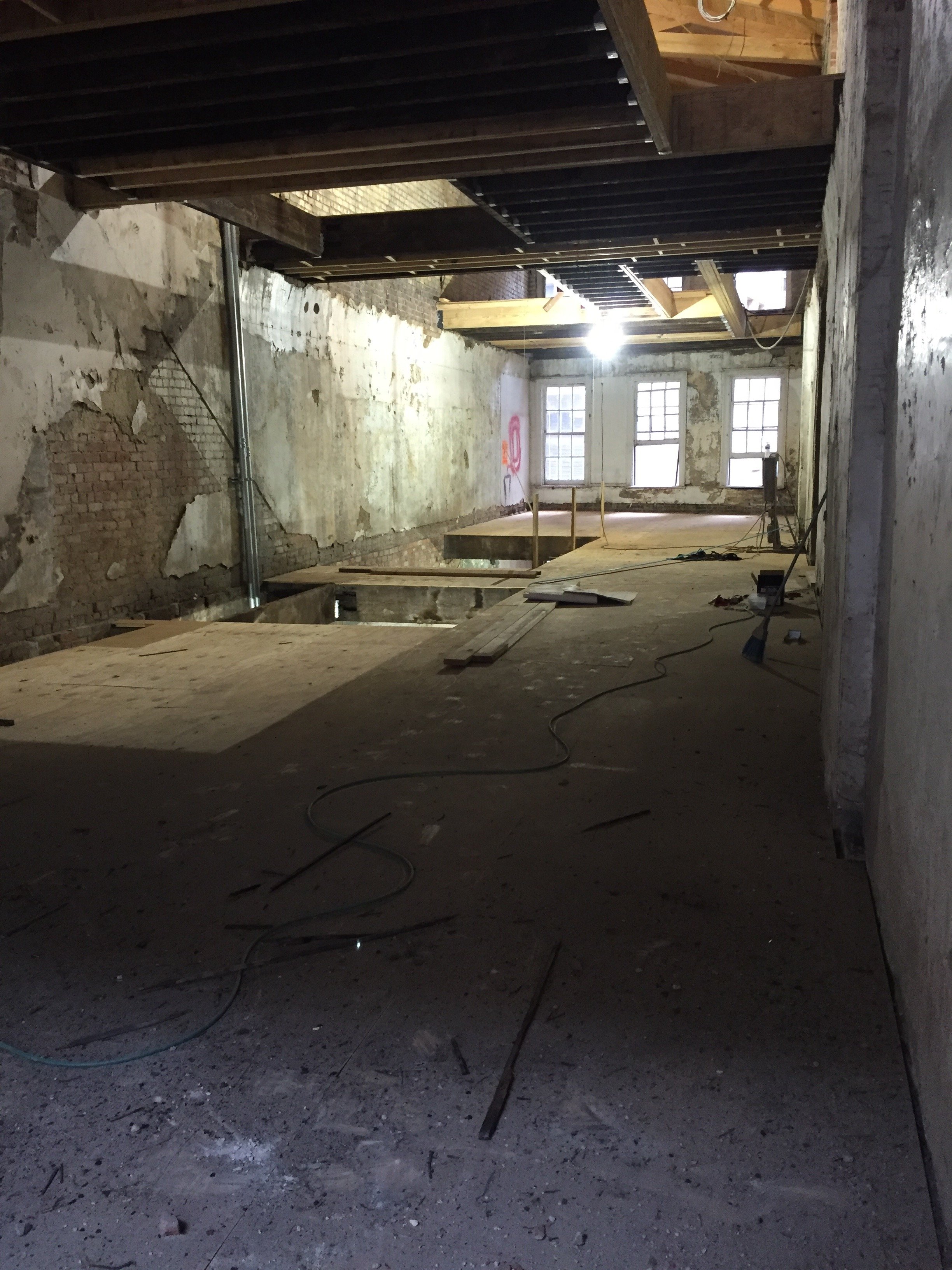 Interior view of a room under renovation, with exposed pipes, electrical wiring, unfinished walls, and construction materials on the floor, illuminated by natural light from multiple windows.