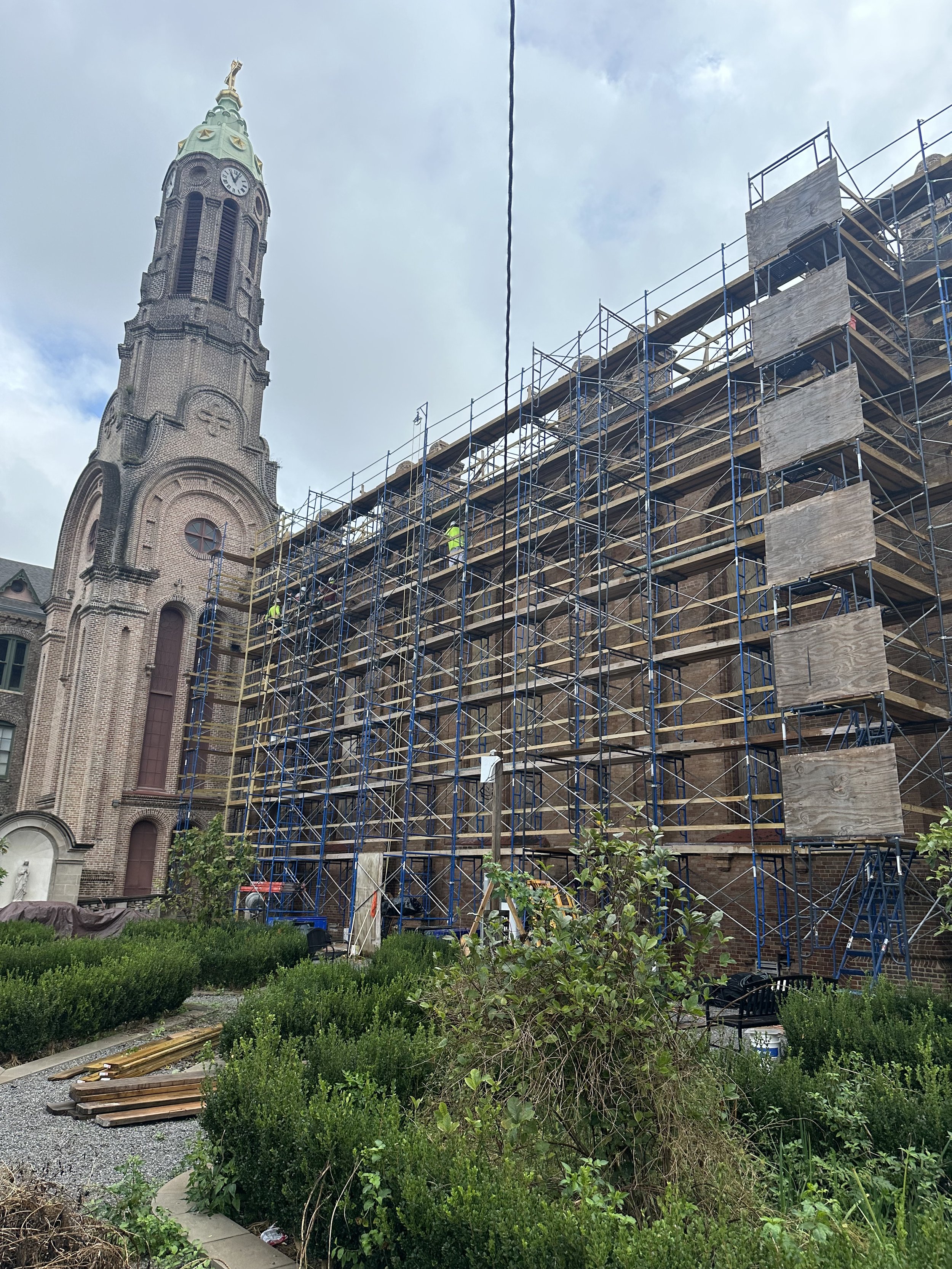 Construction workers working on scaffolding around a brick church with a tall clock tower and a green dome, partly cloudy sky.