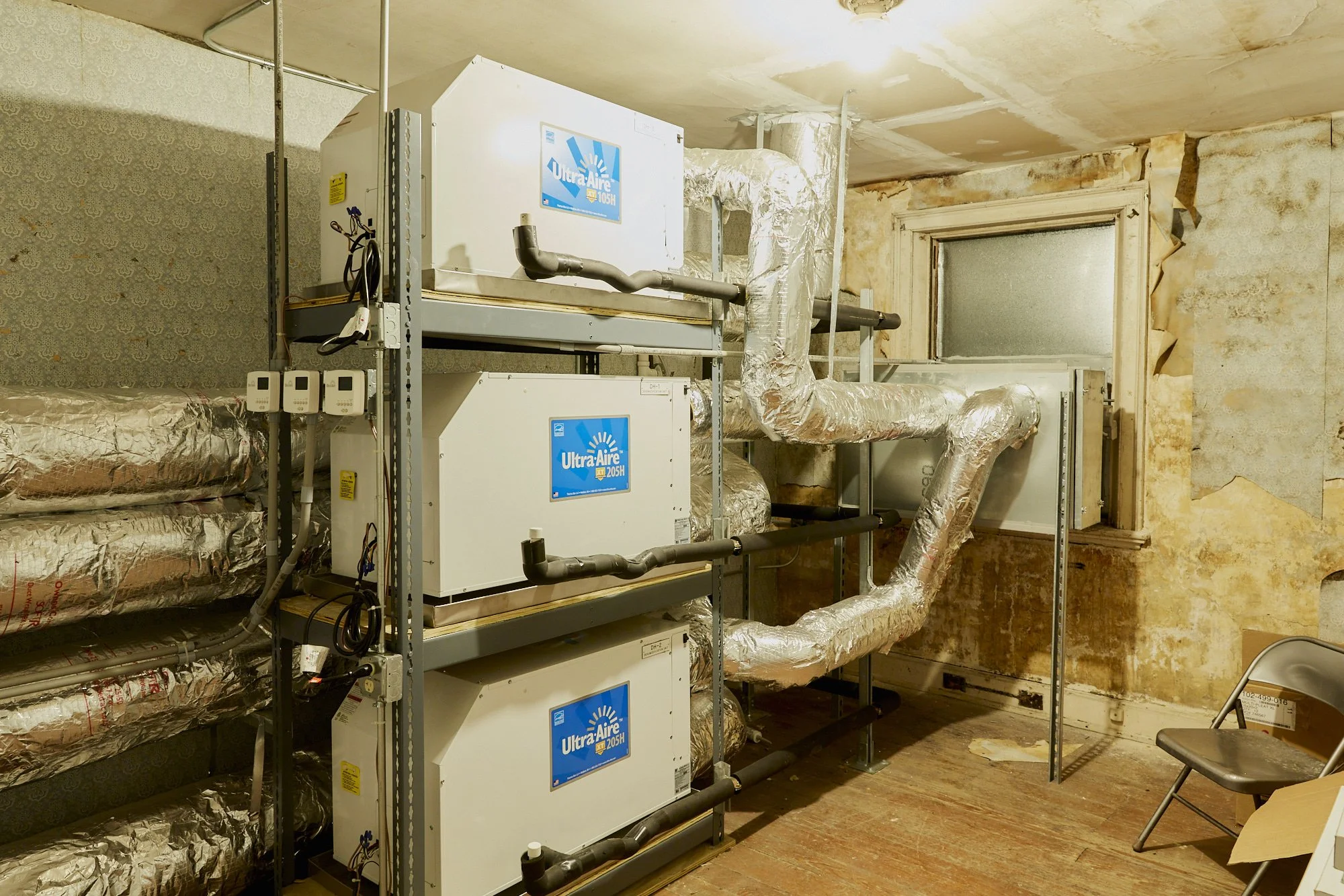 An unfinished room with four HVAC units, insulated and ductwork installed, and a worn wall and window.