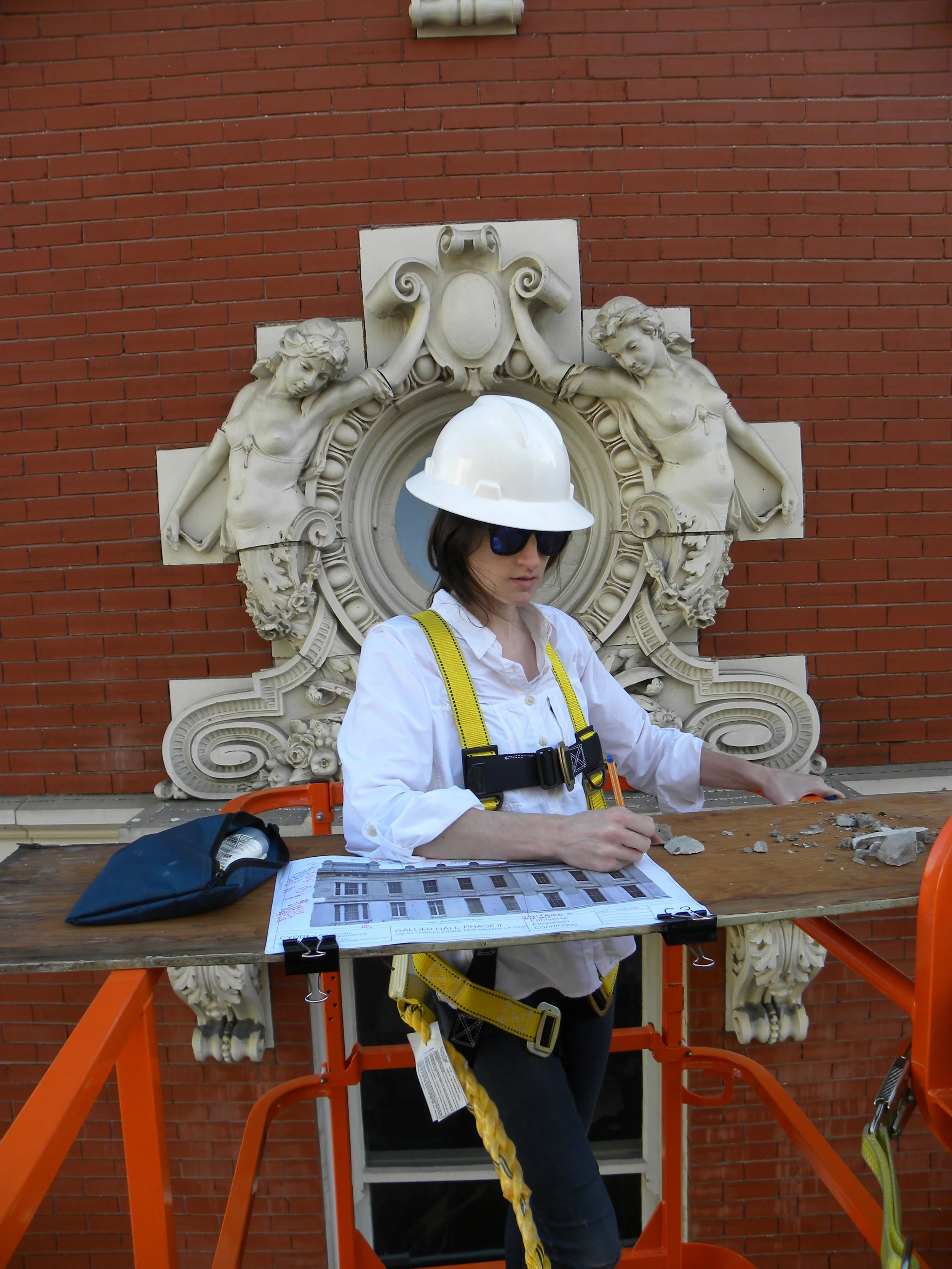 A woman construction worker wearing a white hard hat and sunglasses working at a table on a lift with architectural plans in front of a red brick wall and decorative stone sculptures.
