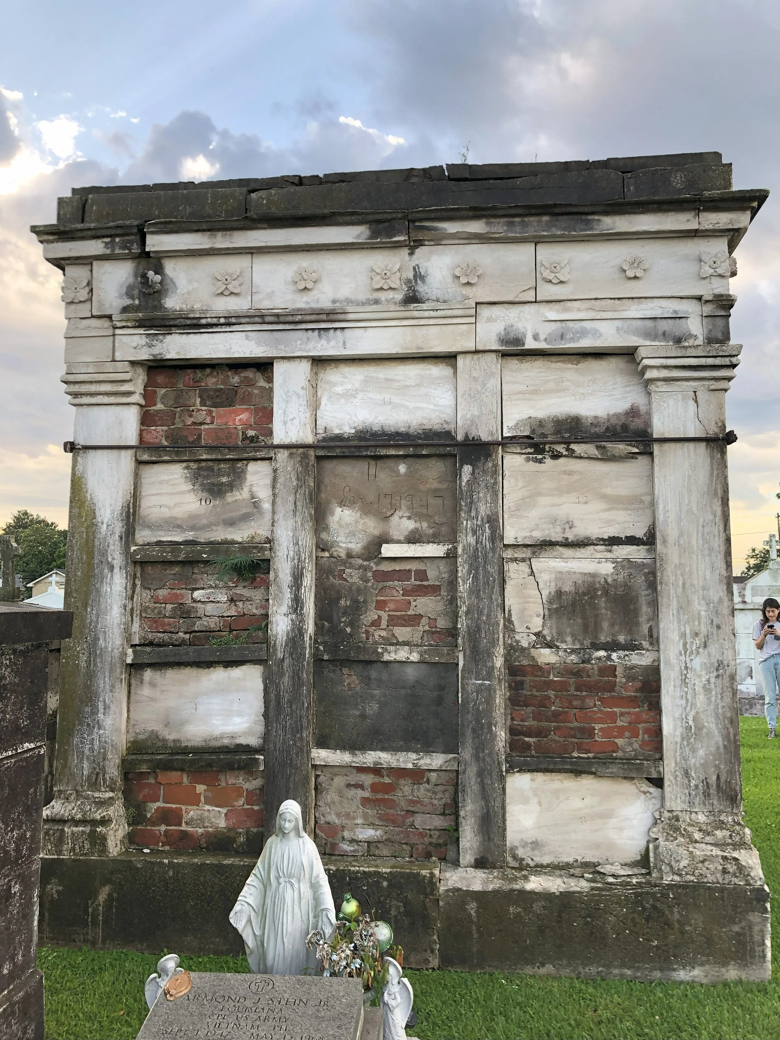 An old, weathered mausoleum with brick and stone construction, showing signs of age and damage, located in a cemetery with a grassy area and a small religious statue in front.