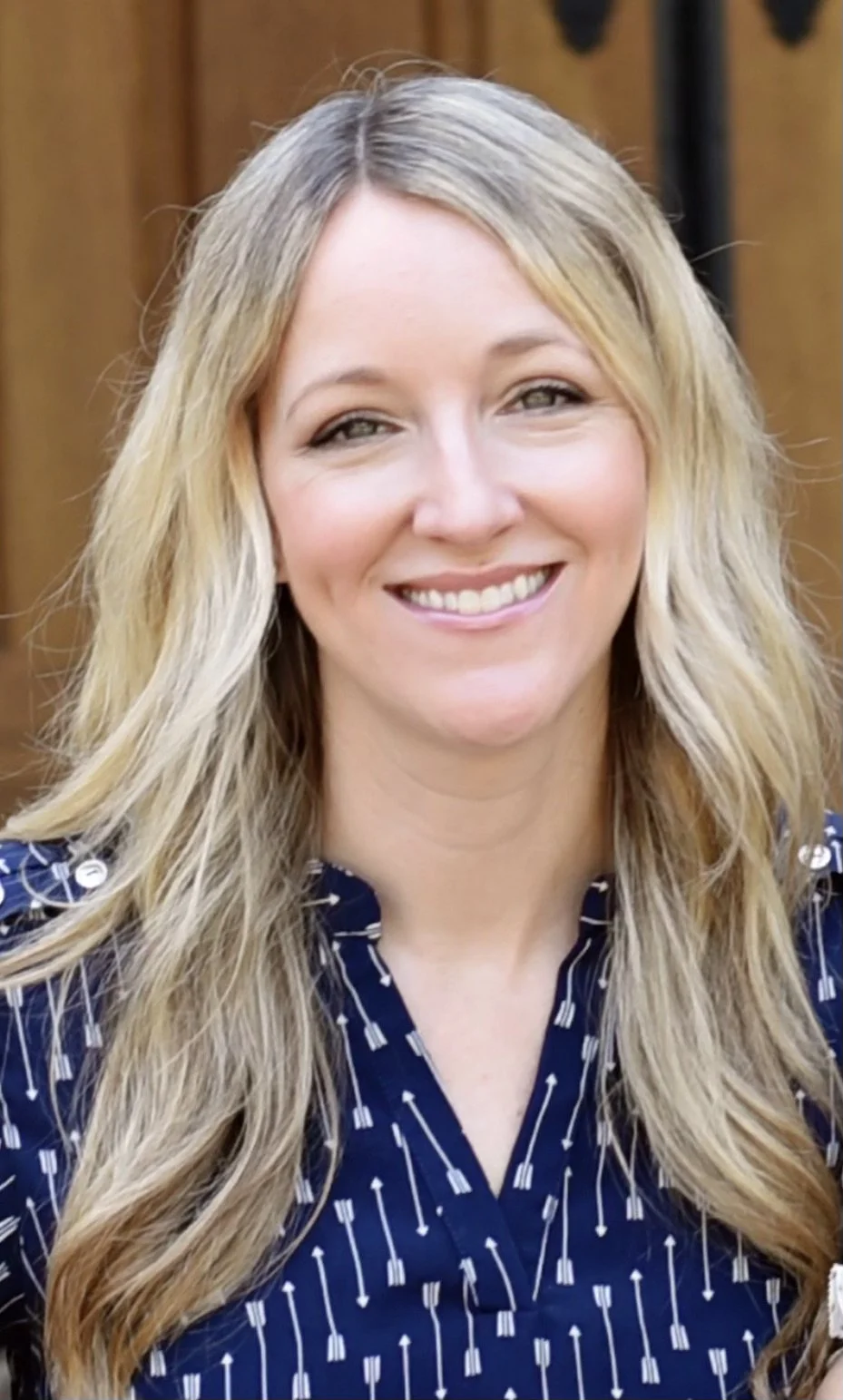 Smiling woman with blonde hair wearing a navy blue blouse with arrow pattern