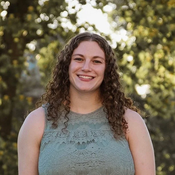 Smiling woman with curly hair wearing a green lace top, standing outdoors.