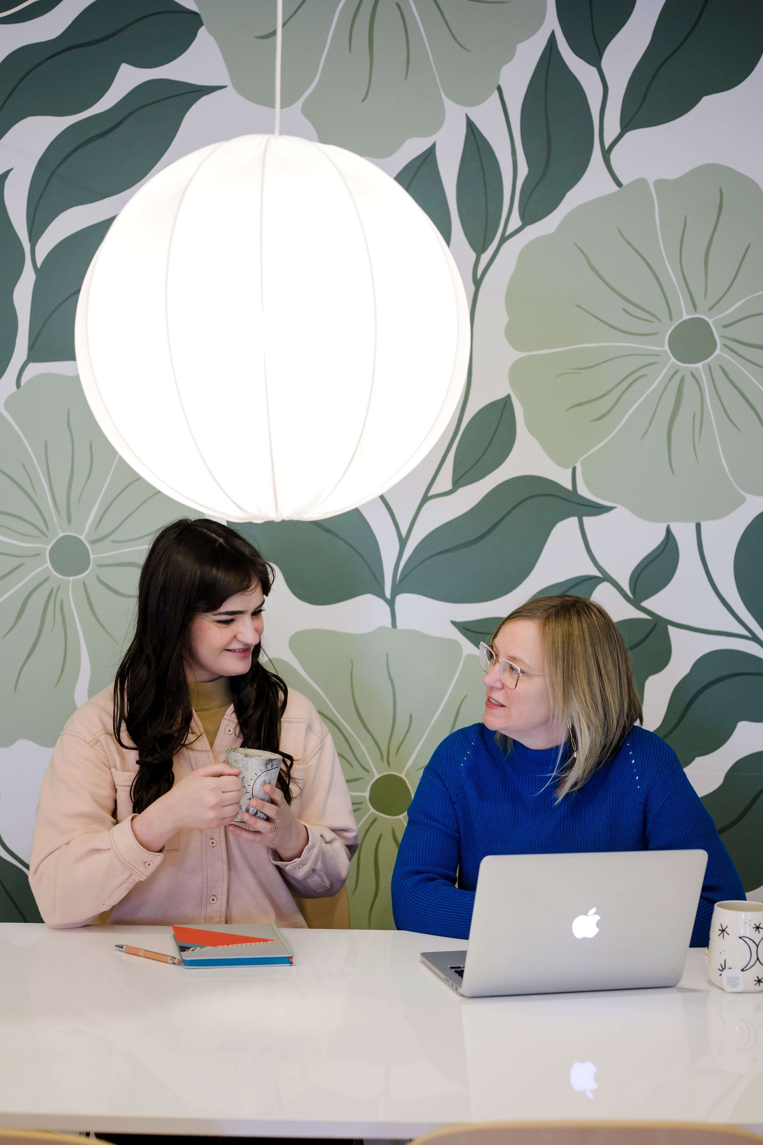 Two women sitting at a white table in an indoor setting with a large white pendant light hanging from the ceiling and a floral patterned wall in the background. One woman with long dark hair and glasses is holding a mug, while the other woman with blonde hair and glasses is working on a silver laptop. Items on the table include a notebook, a pen, a mug with a black star pattern, and a small potted plant.