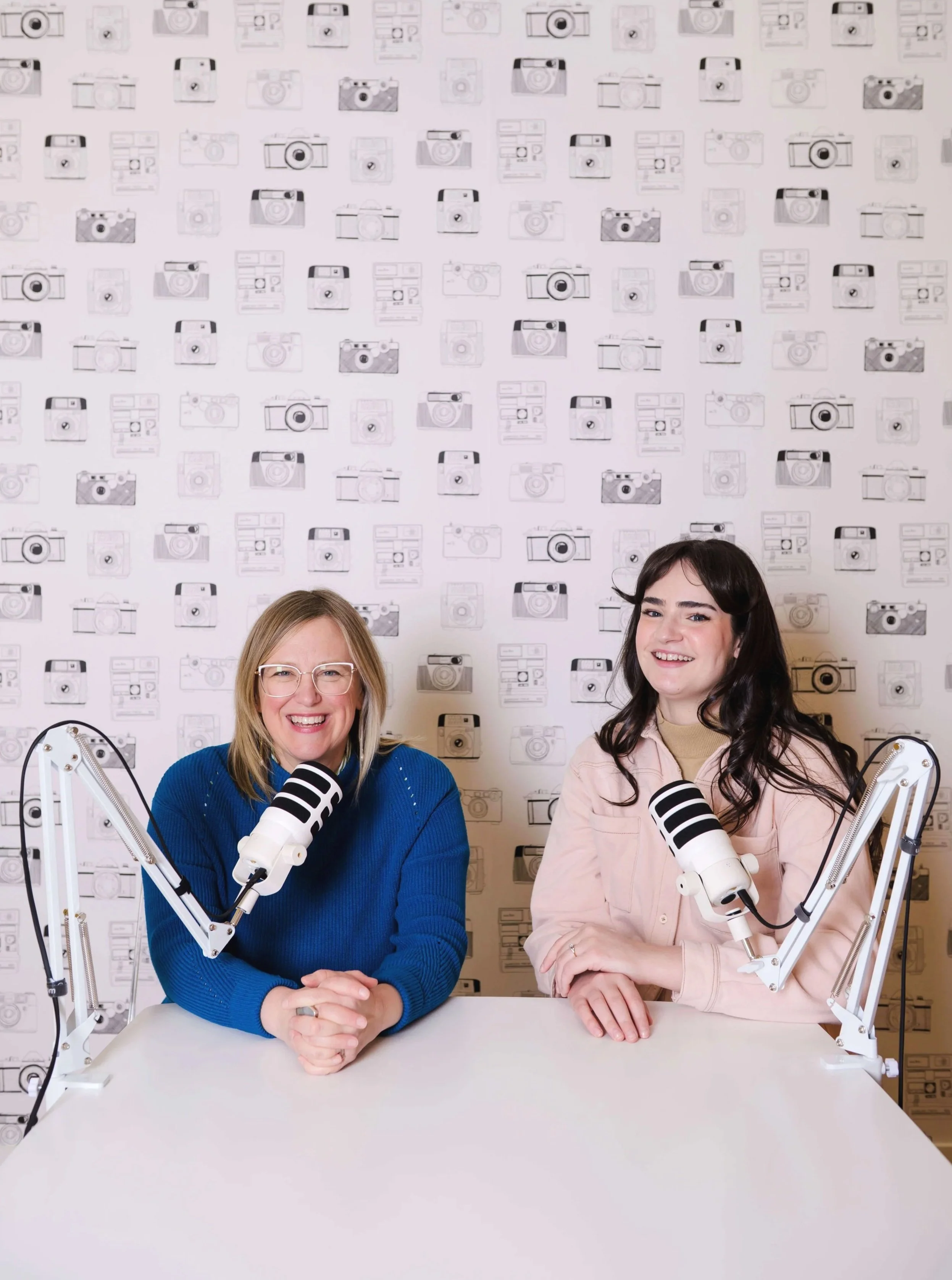Two women sit at a table in a podcast studio with a patterned wallpaper featuring various camera illustrations in the background. They are smiling and wearing microphones mounted on adjustable arms.