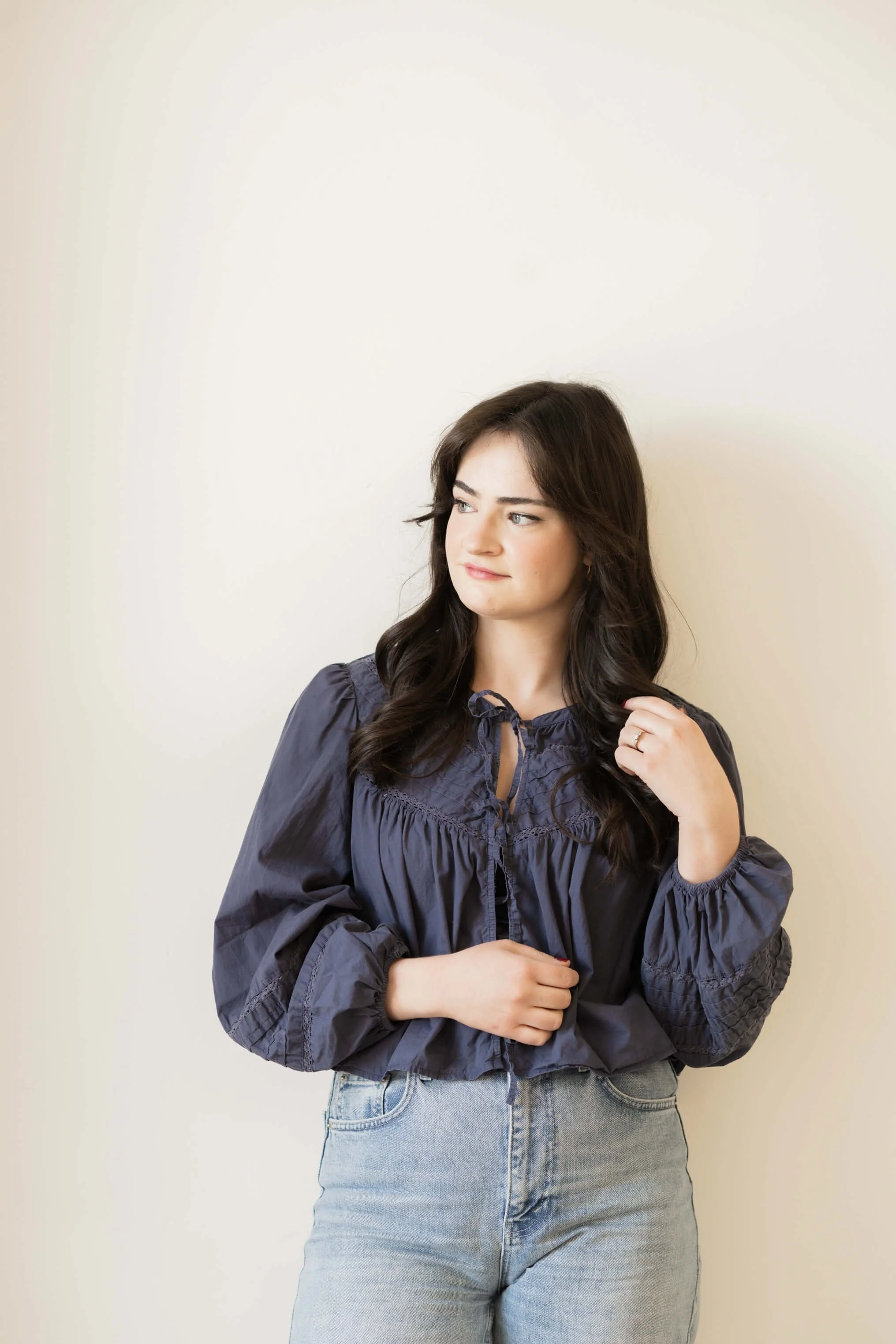 Young woman with long dark hair wearing a navy-blue blouse and light blue jeans, standing against a plain light-colored wall.