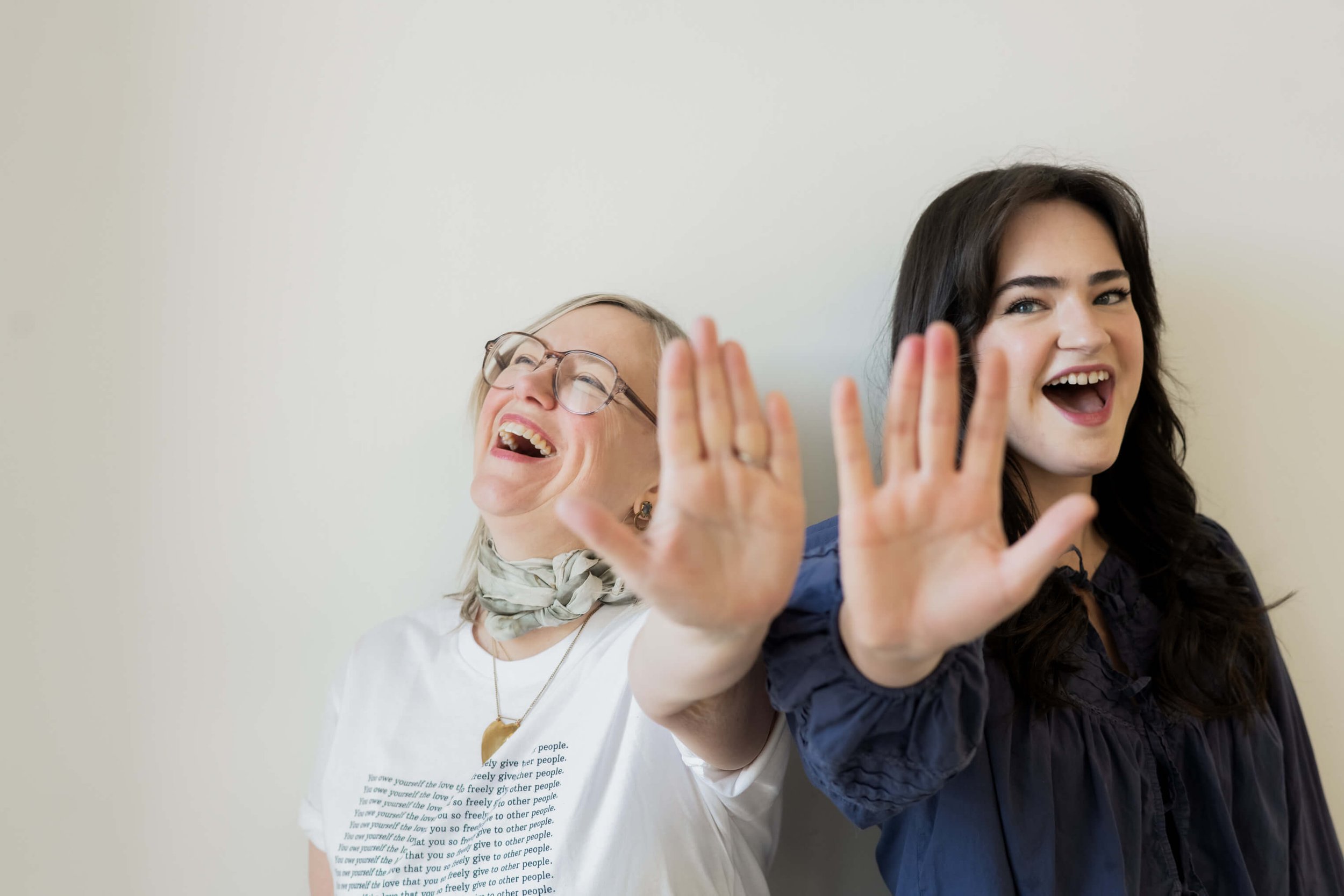 Two women smiling and waving at the camera, standing against a plain white background.