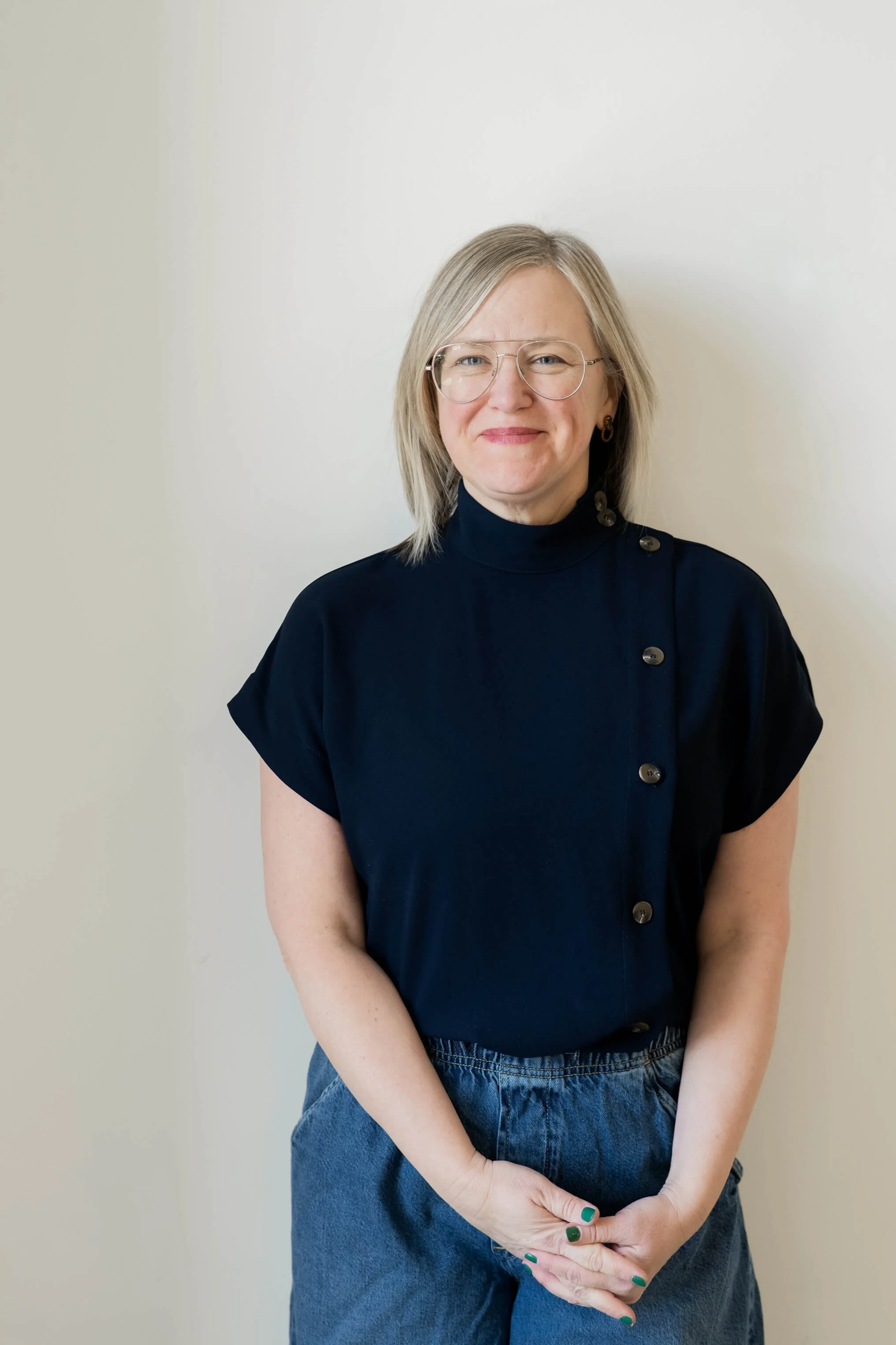 A woman with shoulder-length gray hair, wearing glasses, a navy blue high-neck shirt with buttons on the shoulder, and blue jeans, standing against a plain white wall.