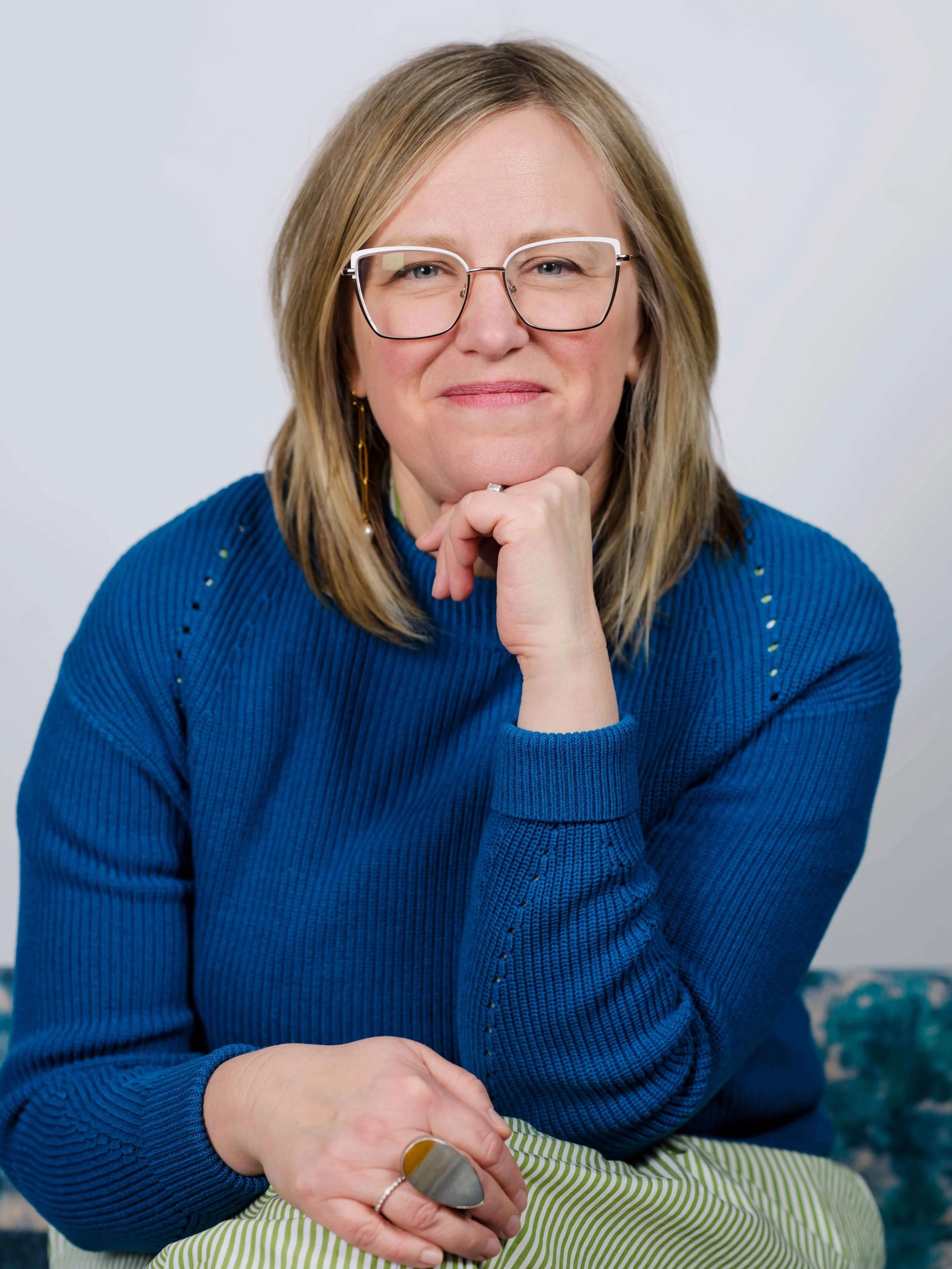 A woman with blond hair and glasses wearing a blue sweater, sitting with her chin resting on her hand, smiling gently at the camera.