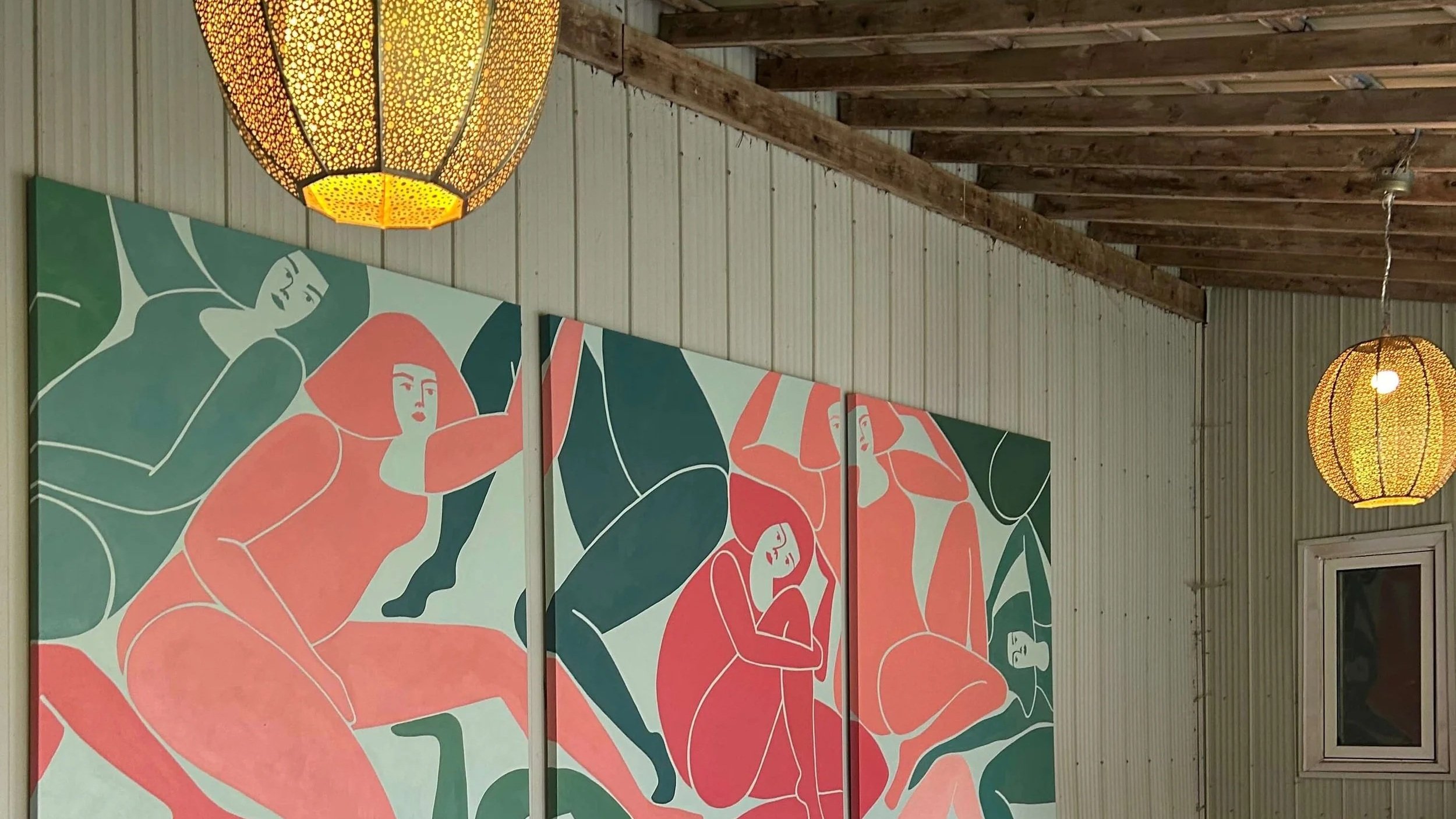 Decorative paintings of abstract human figures and faces on a wall, illuminated by hanging yellow lamps, in a room with a wooden ceiling and beige siding.