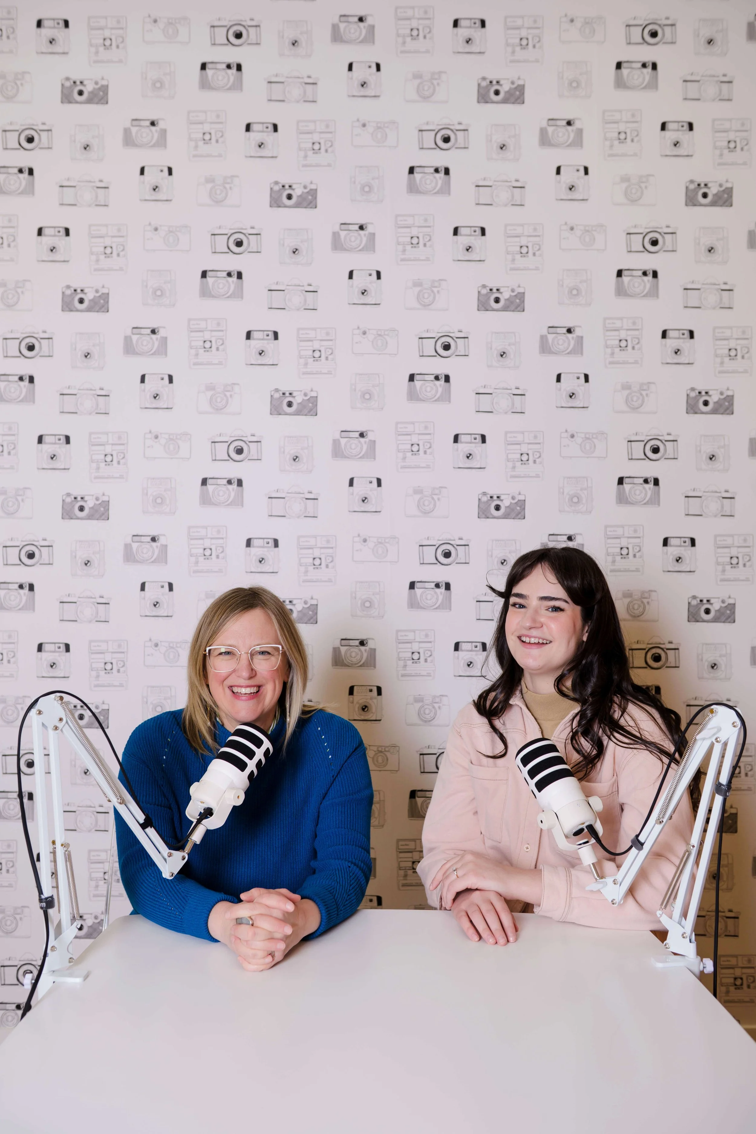 Two women sitting at a table with microphones, smiling, with a backdrop of wallpaper featuring various camera illustrations.