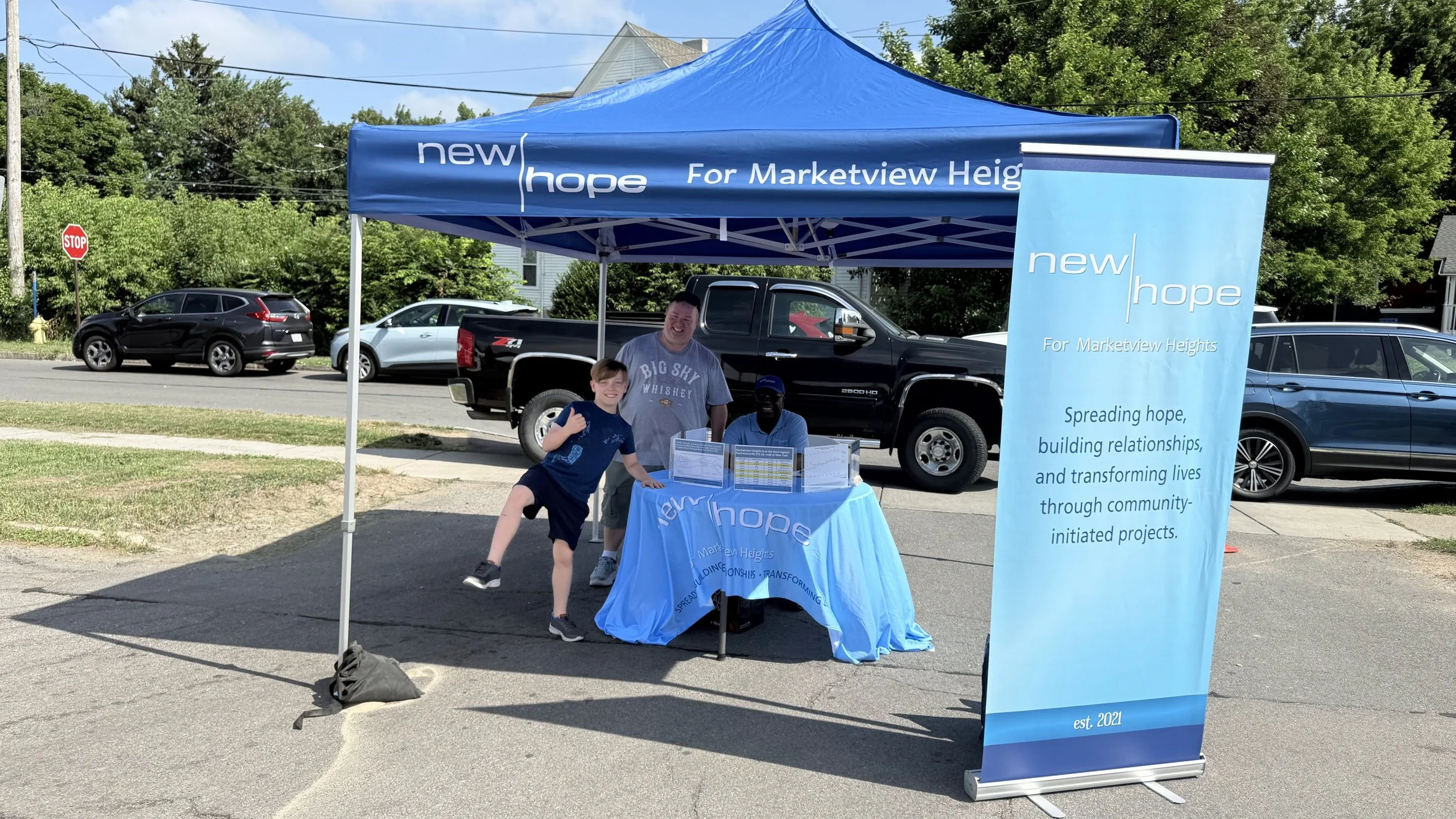 Community outreach booth for 'new hope' organization at Markerview Heights, with a blue canopy tent, a large banner, and three people standing underneath.