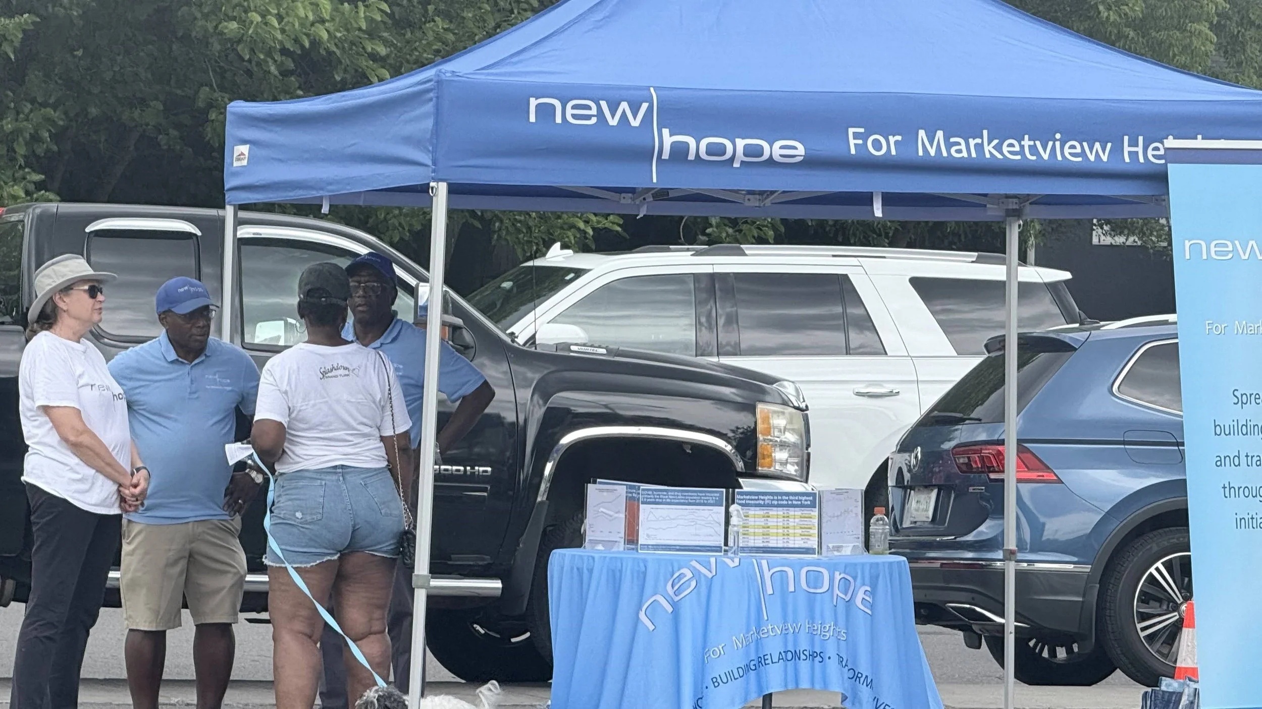 People standing around a booth with a blue canopy that reads 'new hope for Marktwv Heights'. The booth has informational posters and a table with a blue tablecloth, set up in a parking lot. Several cars are parked behind the booth, and trees are in t