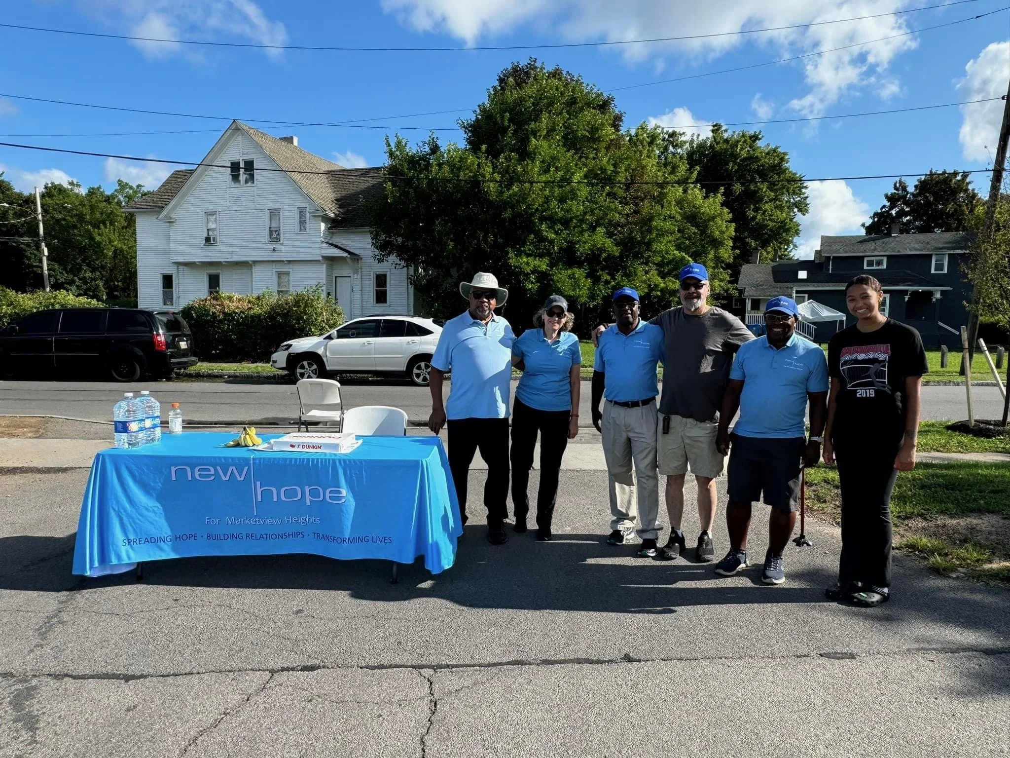 Group of six people standing behind a blue table with bottled water, bananas, and a sign that reads "new hope for Marketview Heights." They are outdoors on a sunny day with houses and cars in the background.