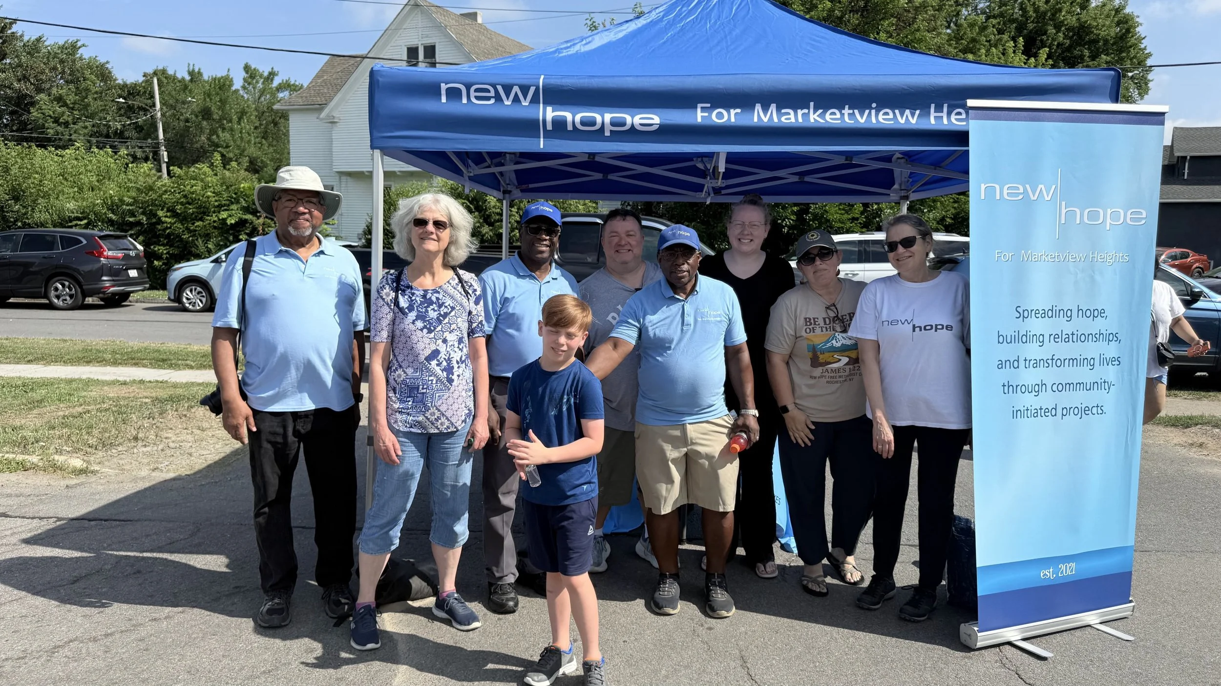 Group of diverse people standing under a blue tent that says "new hope" and a matching banner, with a residential street in the background. The group is smiling and posing for the photo.