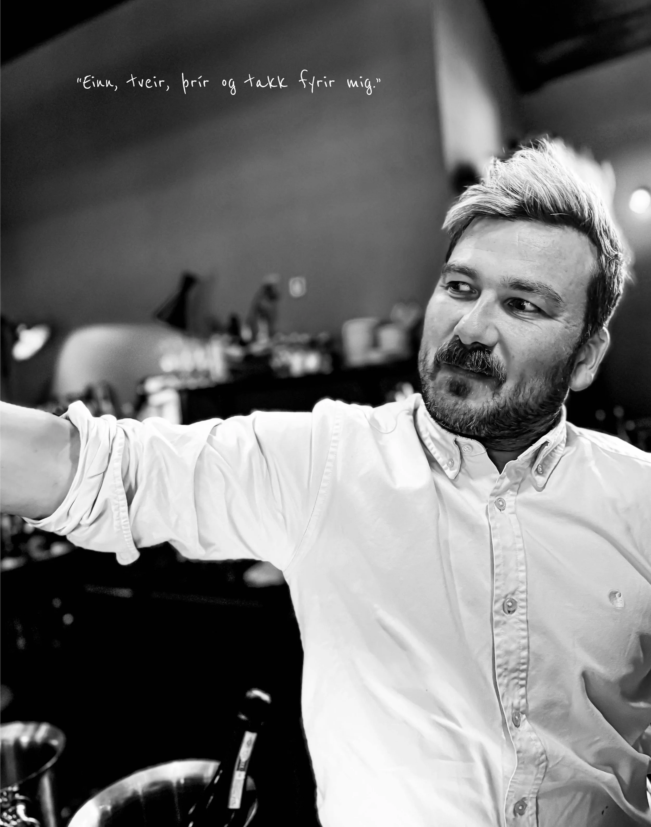 Black and white photo of a man with short, light-colored hair and a beard, wearing a light-colored button-up shirt. He is looking off to the side, with some blurred objects and a quote in Icelandic in the background.