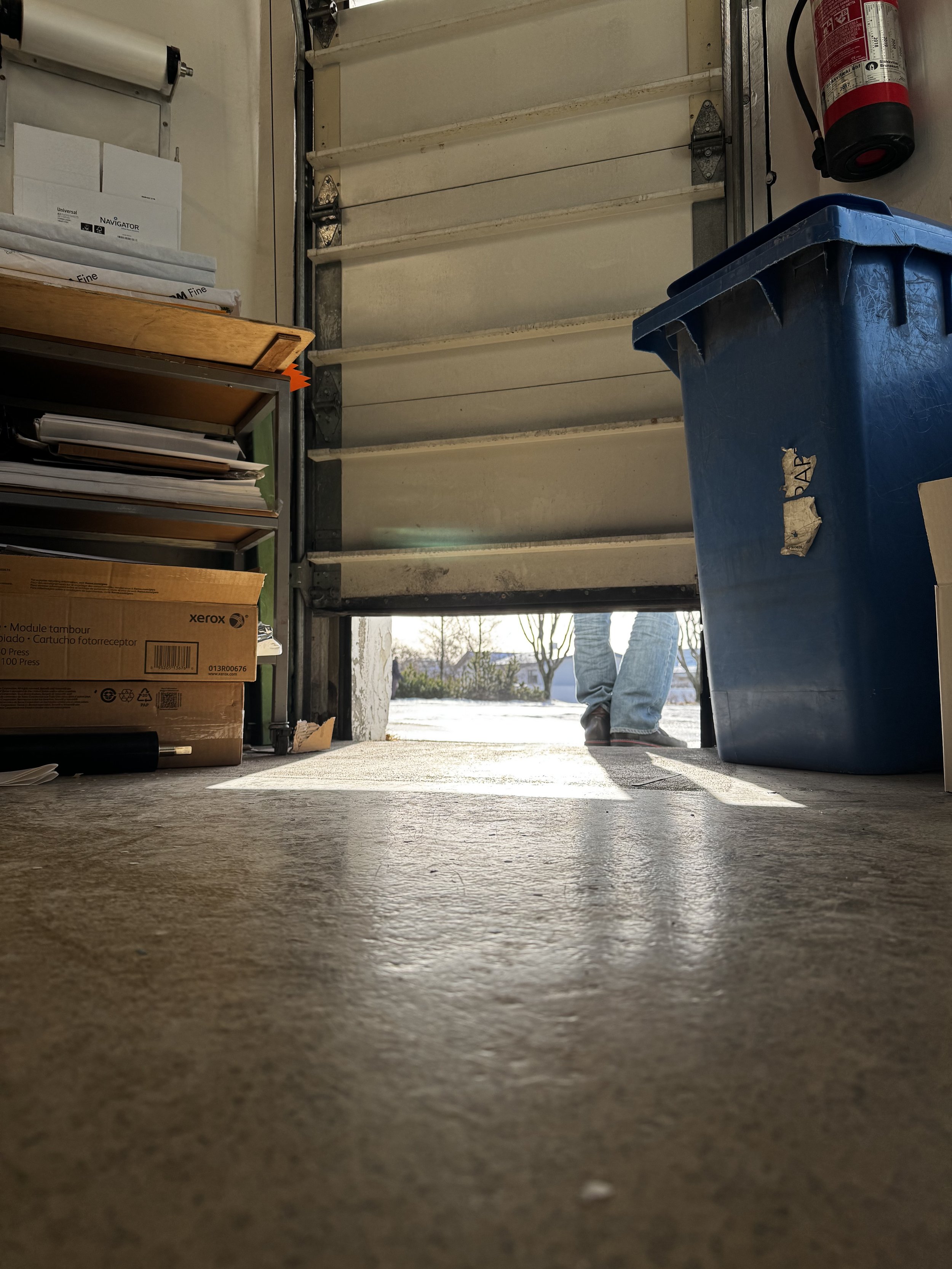 Looking from the garage floor towards the garage door, with a person's legs visible outside the garage. Various items are stored on shelves and in bins inside the garage, including a fire extinguisher on the wall.