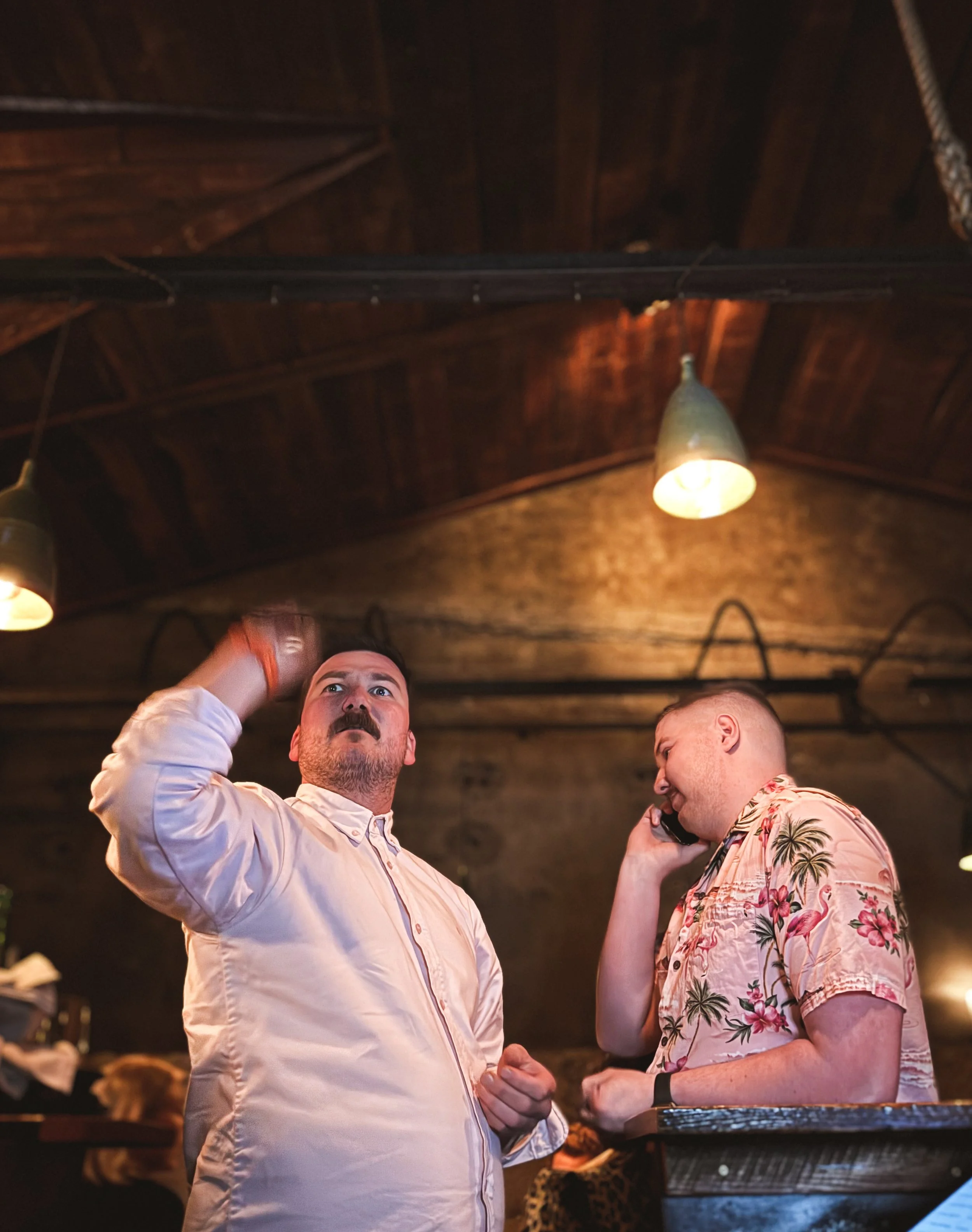 Two men are in a restaurant, one holding a phone to his ear and the other adjusting his hair. The man on the left is wearing a light-colored shirt and the man on the right is wearing a tropical shirt with pink and green floral patterns. The ceiling is dark wood with lights hanging from it.