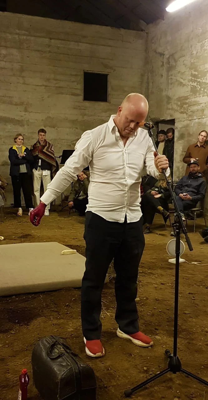 A man wearing a white shirt, black pants, and red shoes, stands in front of a microphone in a room with concrete walls, surrounded by an audience.