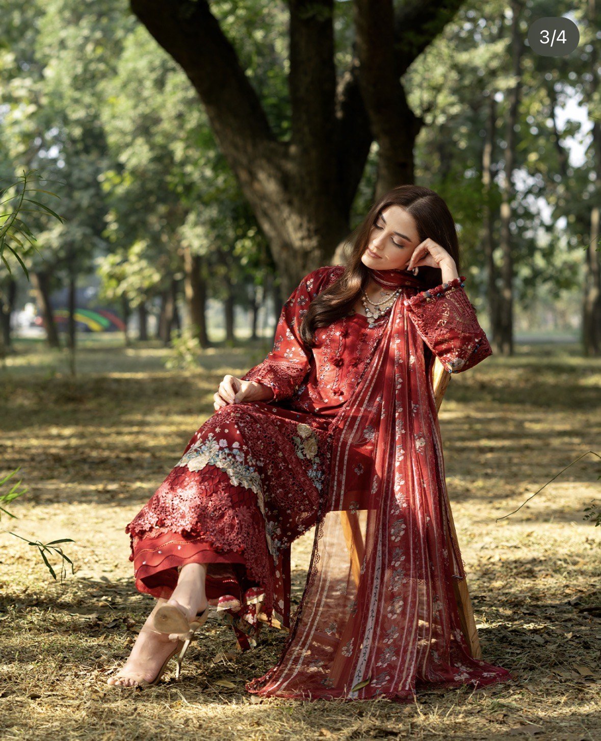 A woman dressed in a red traditional outfit sitting on a chair in a park with trees in the background, resting her head on her hand with her eyes closed.