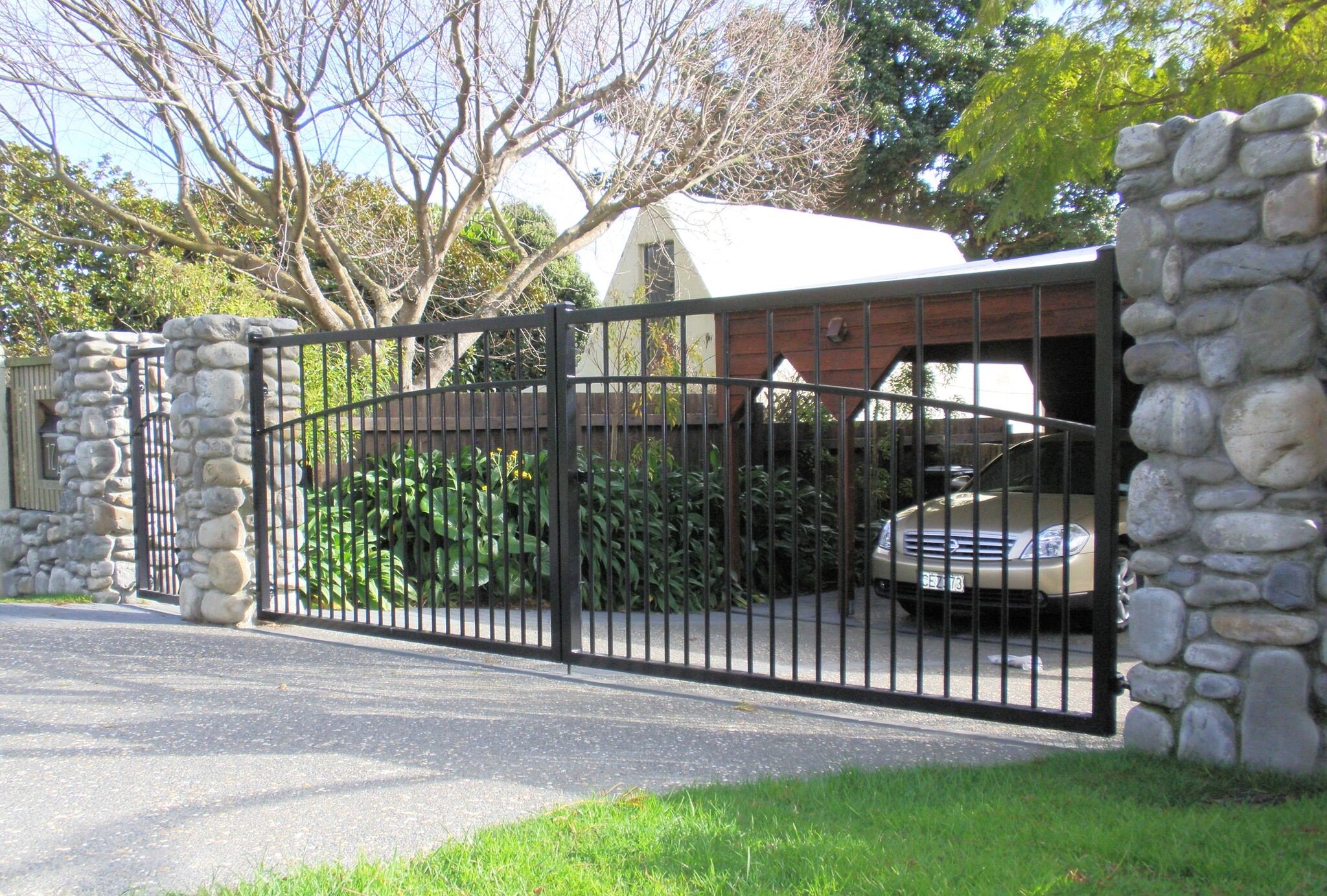 Metal driveway gate with stone pillars, car visible behind in a garage, surrounded by trees and greenery.
