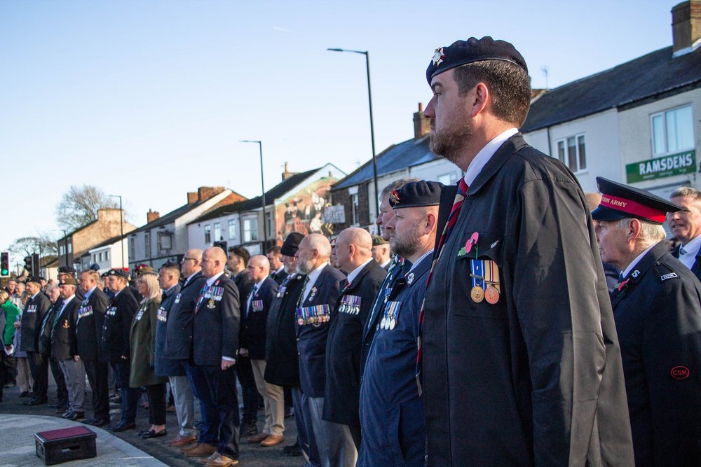 A group of men, some veterans, standing in a line during a memorial or remembrance event. Many are wearing military-style medals and remembrance poppies.