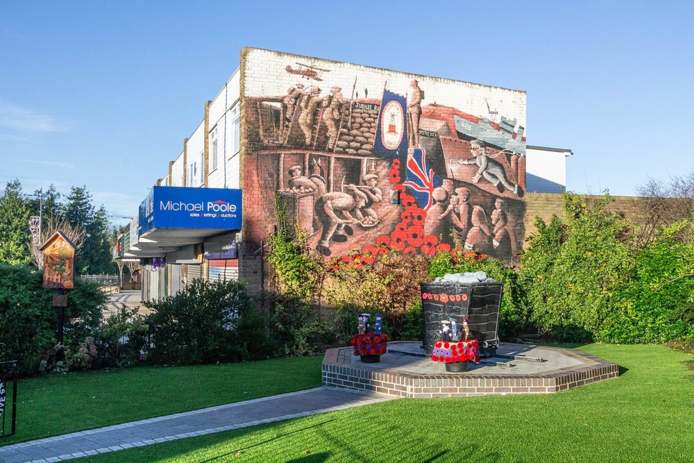 Street mural on a brick building depicting soldiers, a plane, and a Union Jack flag with a clock, with flowers and bushes in the foreground.