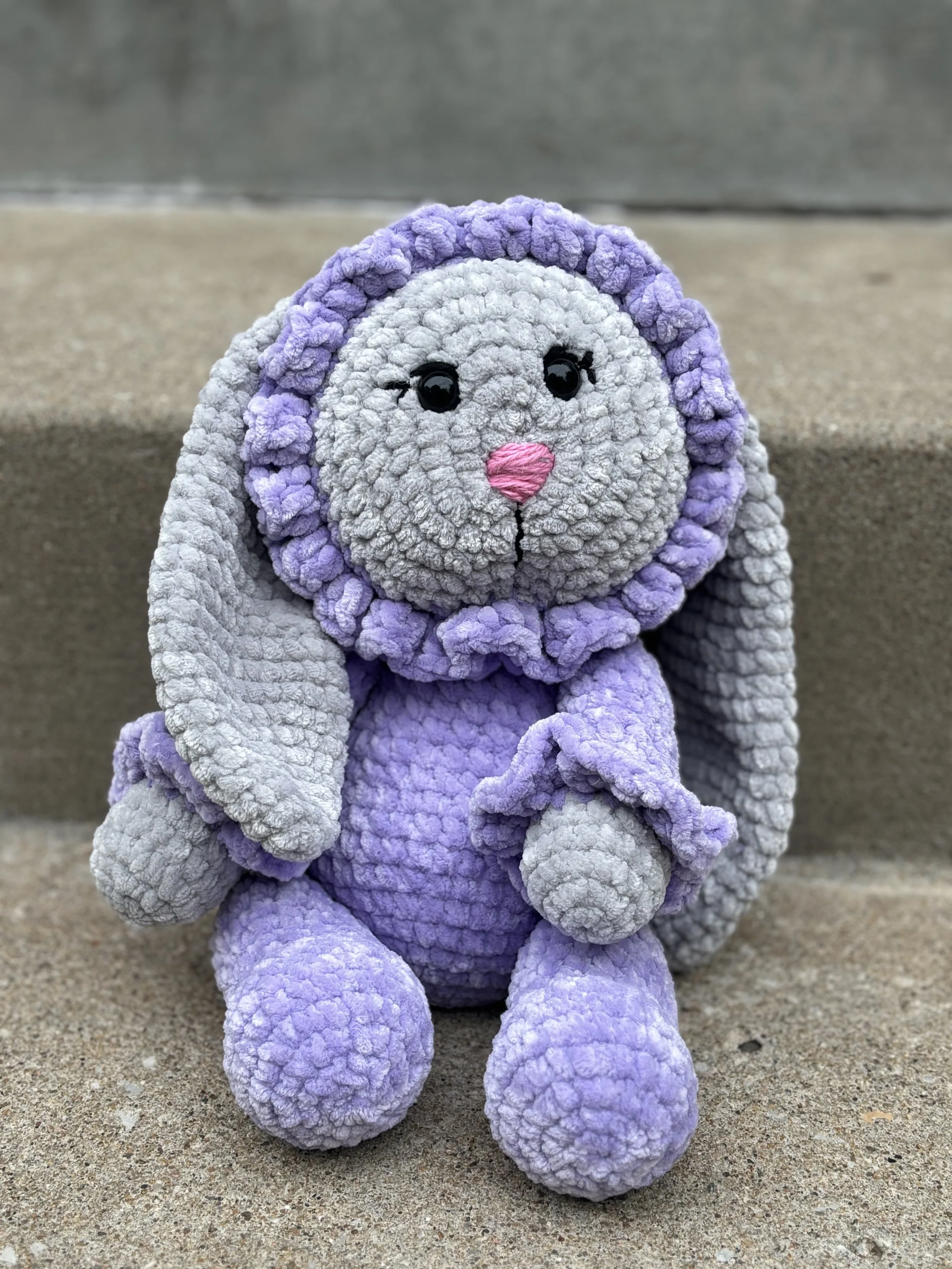 A plush toy of a bunny with purple and gray textured fabric, black eyes, and pink nose sitting on concrete steps.
