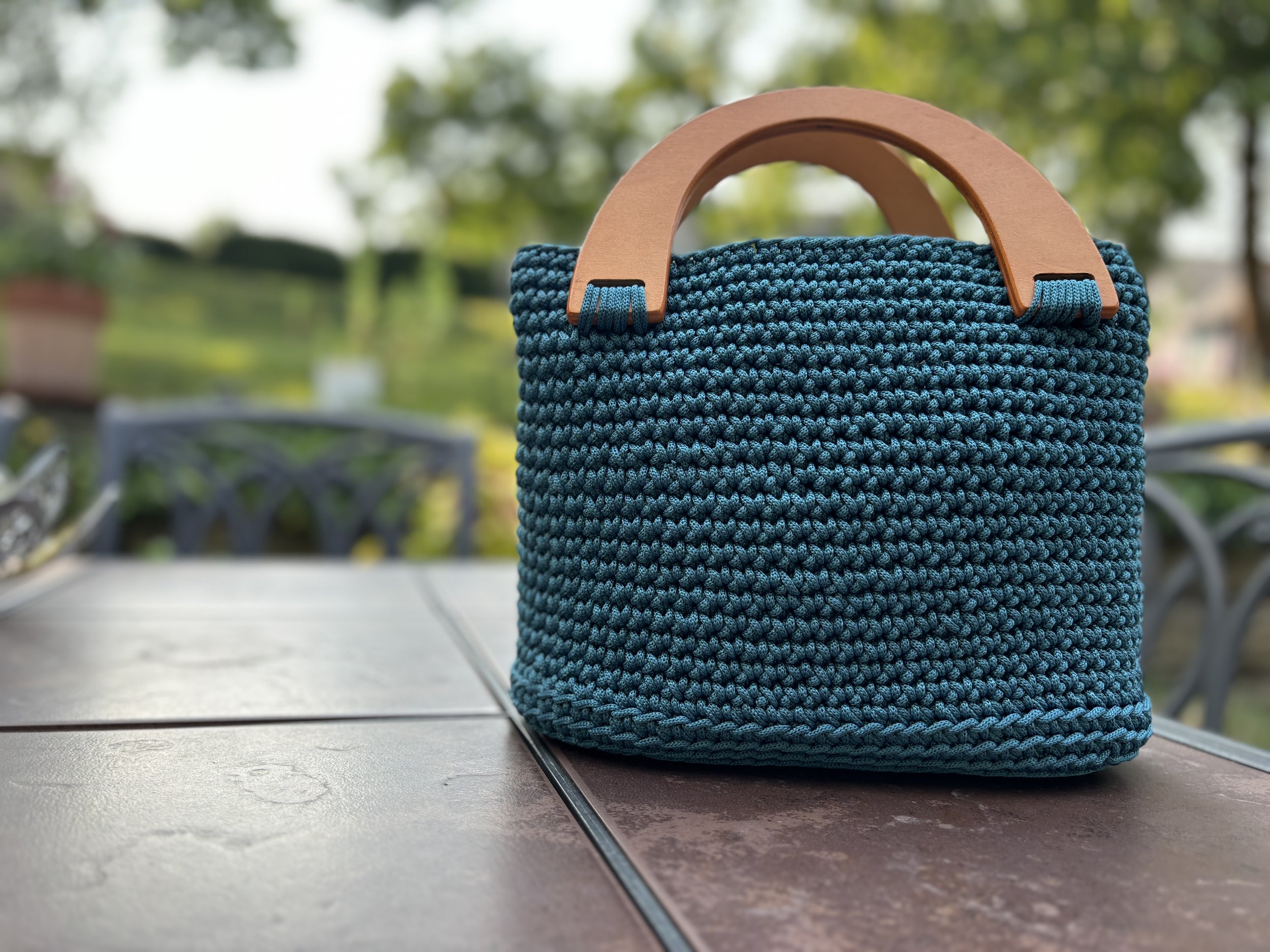 Blue woven handbag with wooden handles placed on an outdoor table in a garden setting.