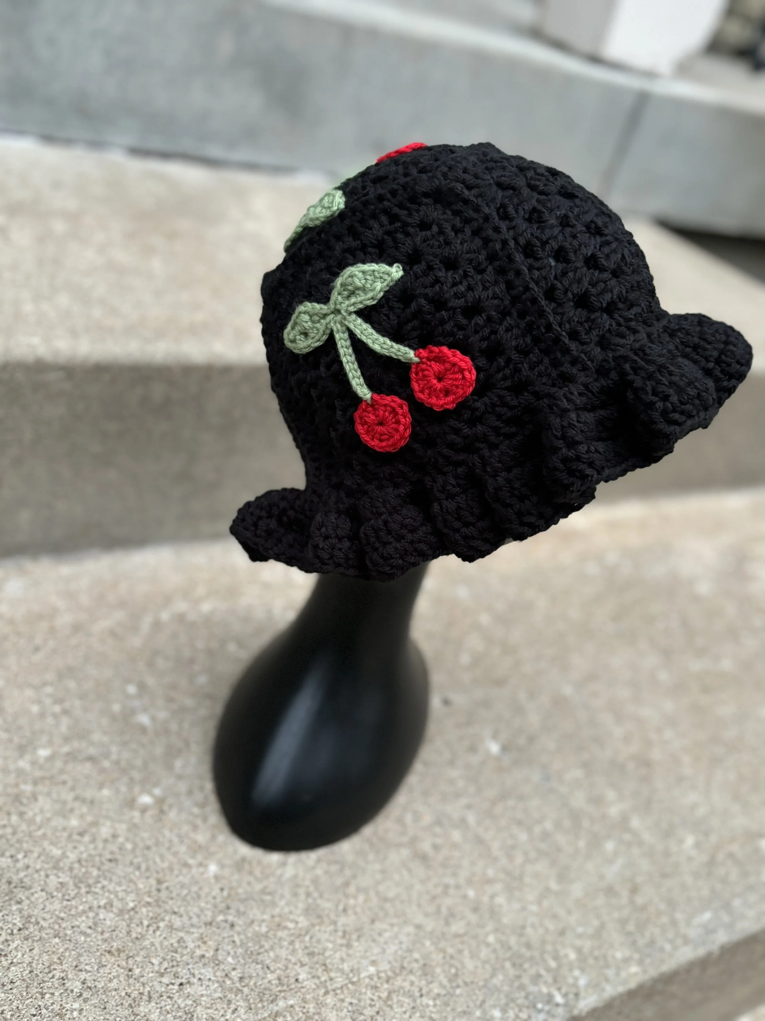 Black crochet hat with red cherry designs on mannequin; placed on concrete steps.