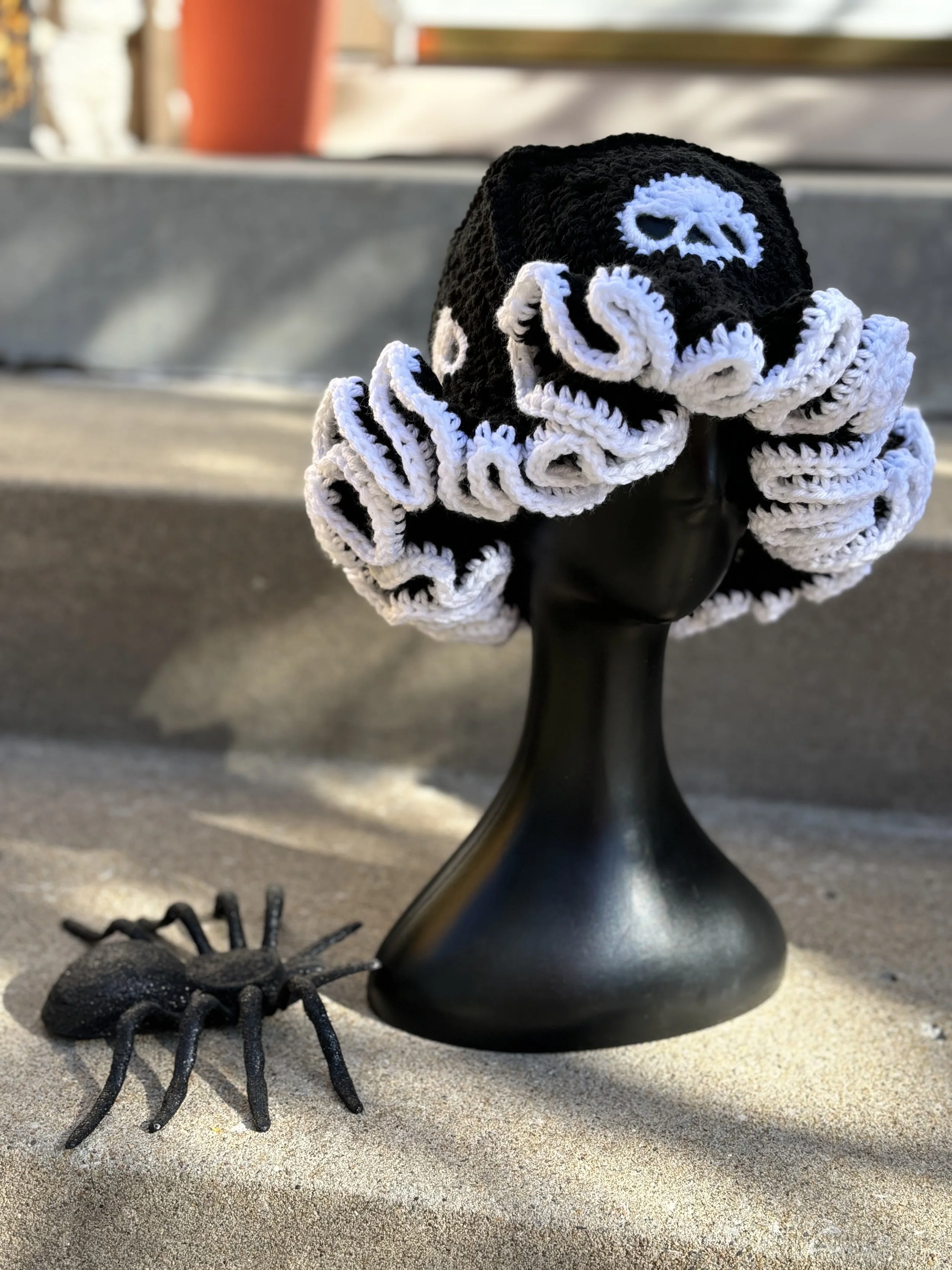 A black mannequin head displays a black and white crocheted hat with a skull design, next to a black decorative spider on a concrete step.
